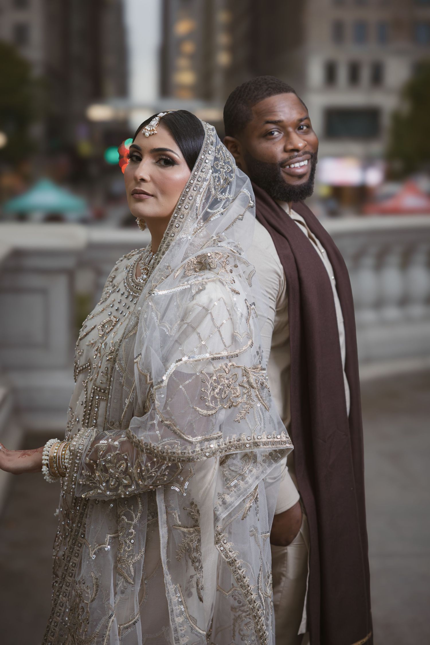 Interracial couple in traditional Islamic wedding attire posing back-to-back on urban city bridge.