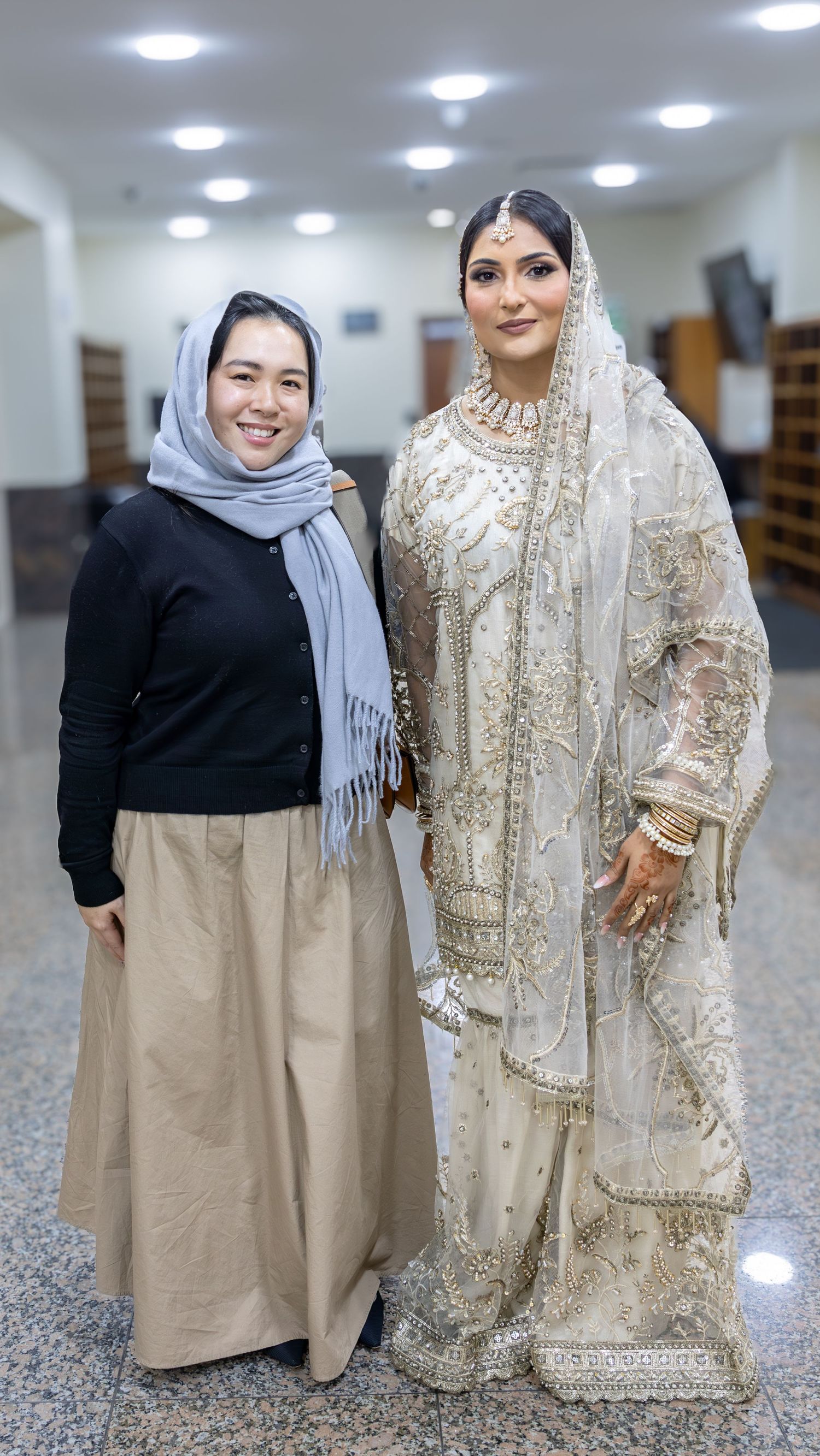 A smiling woman in gray hijab stands beside a bride wearing elegant white embroidered South Asian bridal dress.