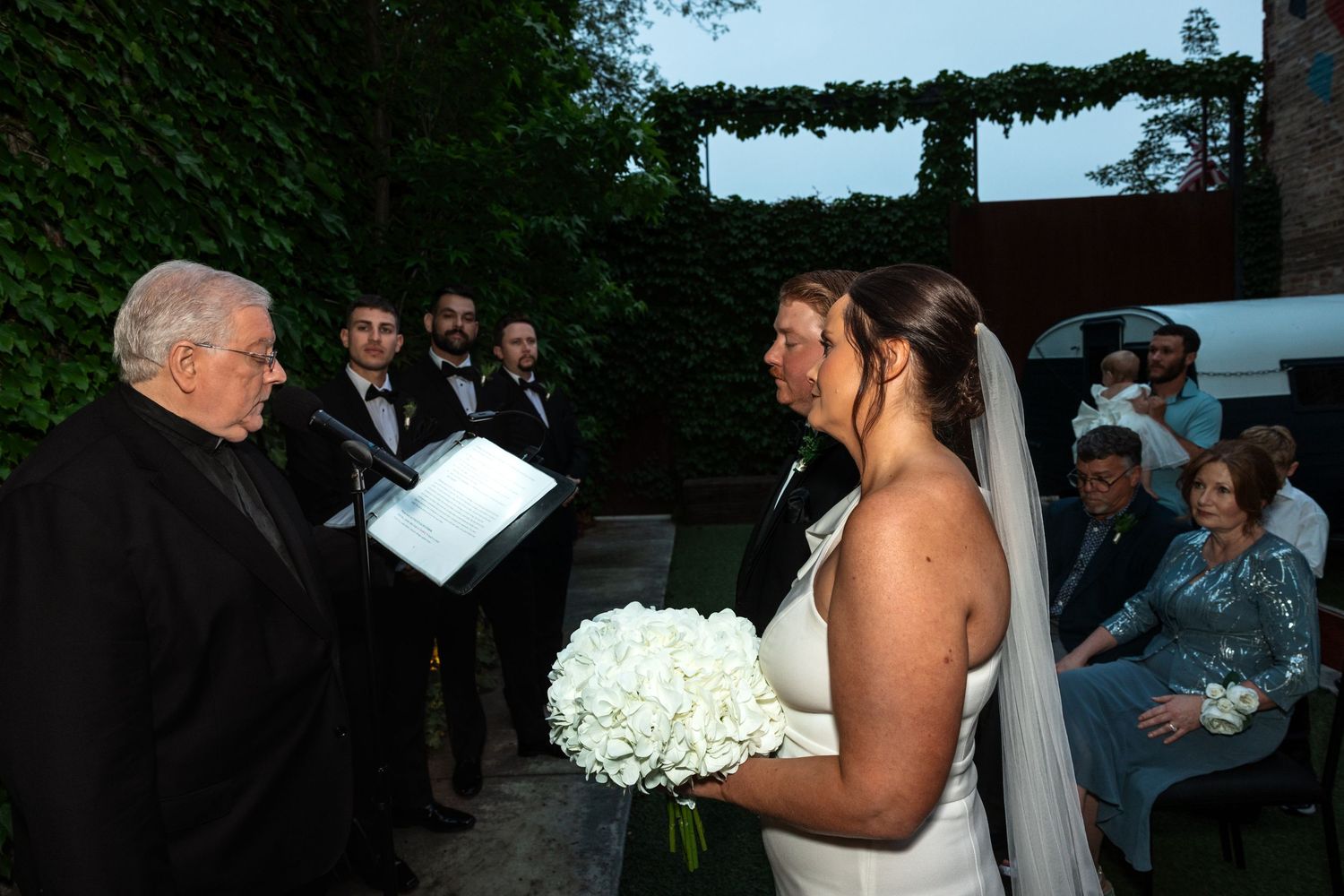 Bride holds white bouquet while officiant reads vows during an outdoor wedding ceremony at dusk.