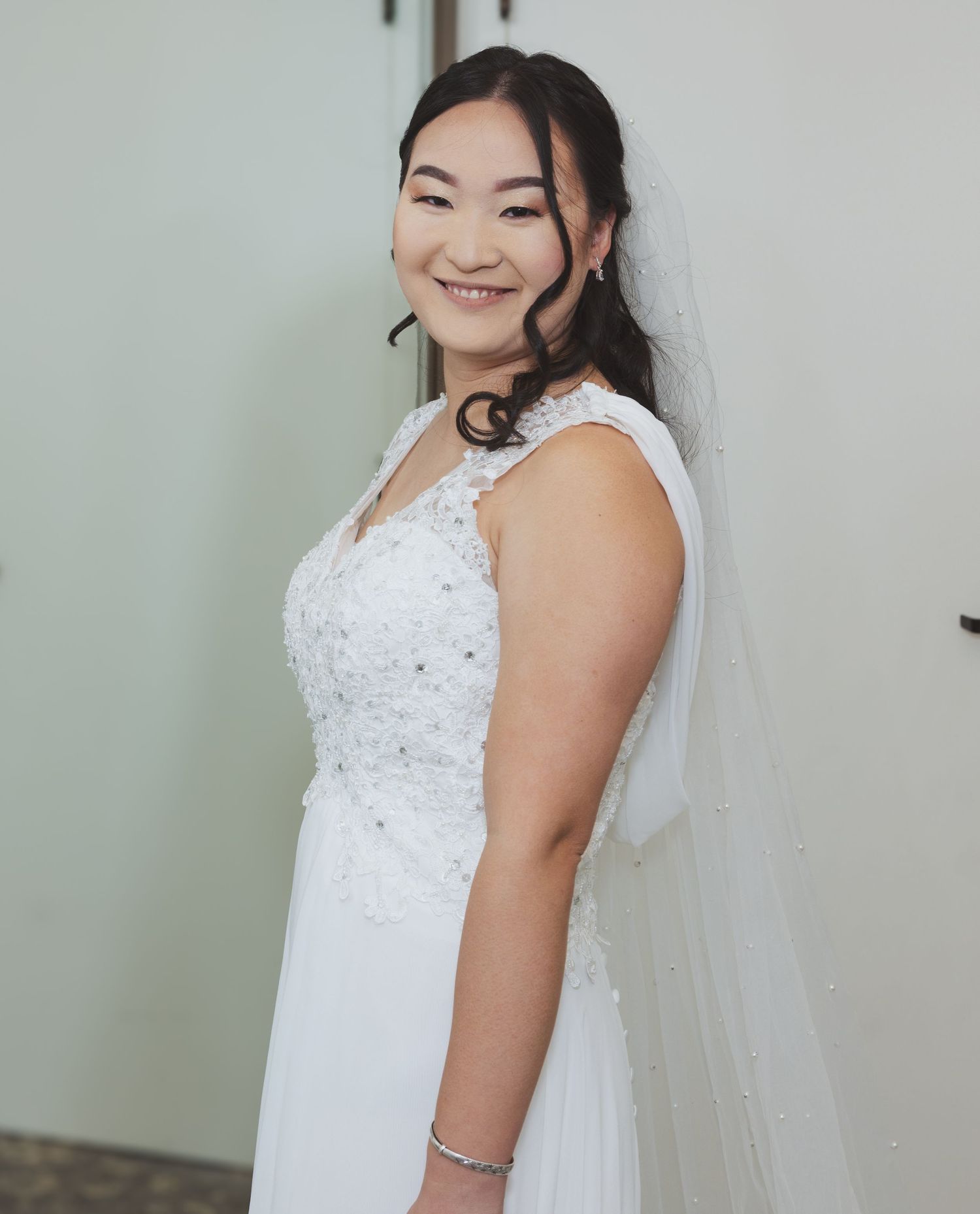 Smiling bride in a lace beaded white wedding gown with veil and bracelet posing elegantly.