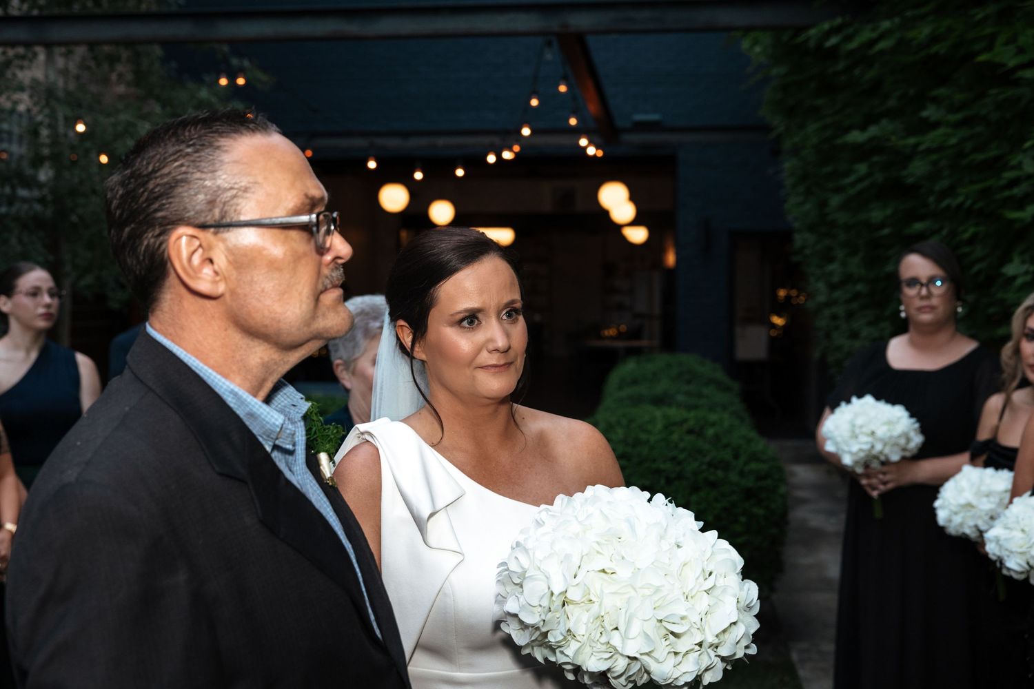 Bride in white gown with veil holding white floral bouquet being escorted at outdoor evening wedding.