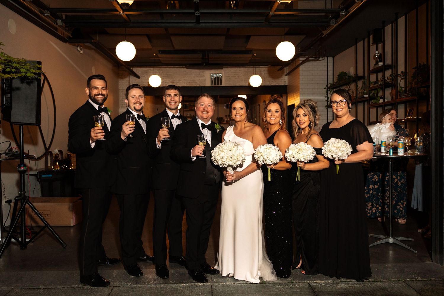 Wedding party in black attire with bride in white gown holding white floral bouquets celebrating with champagne.
