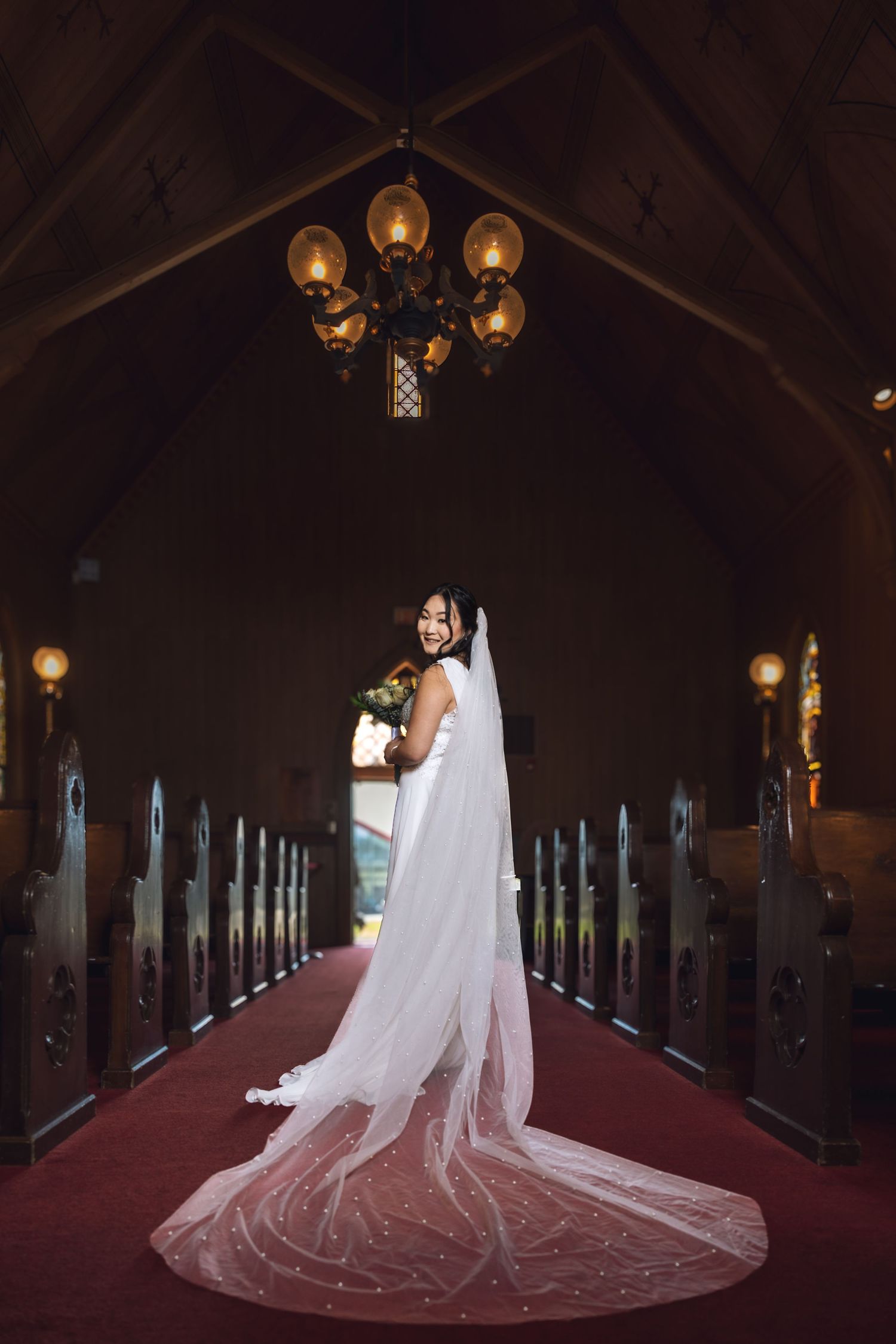 Radiant bride in white dress with flowing cathedral veil posing in red-carpeted church aisle.