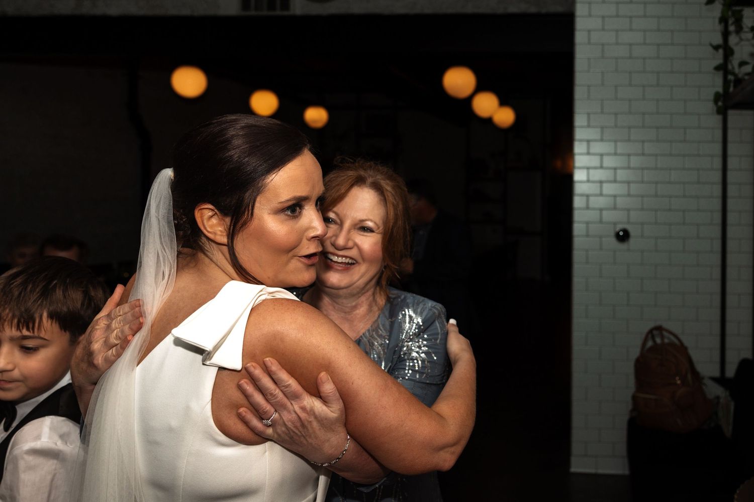 Bride in white wedding dress receives warm hug from older woman guest at wedding reception.