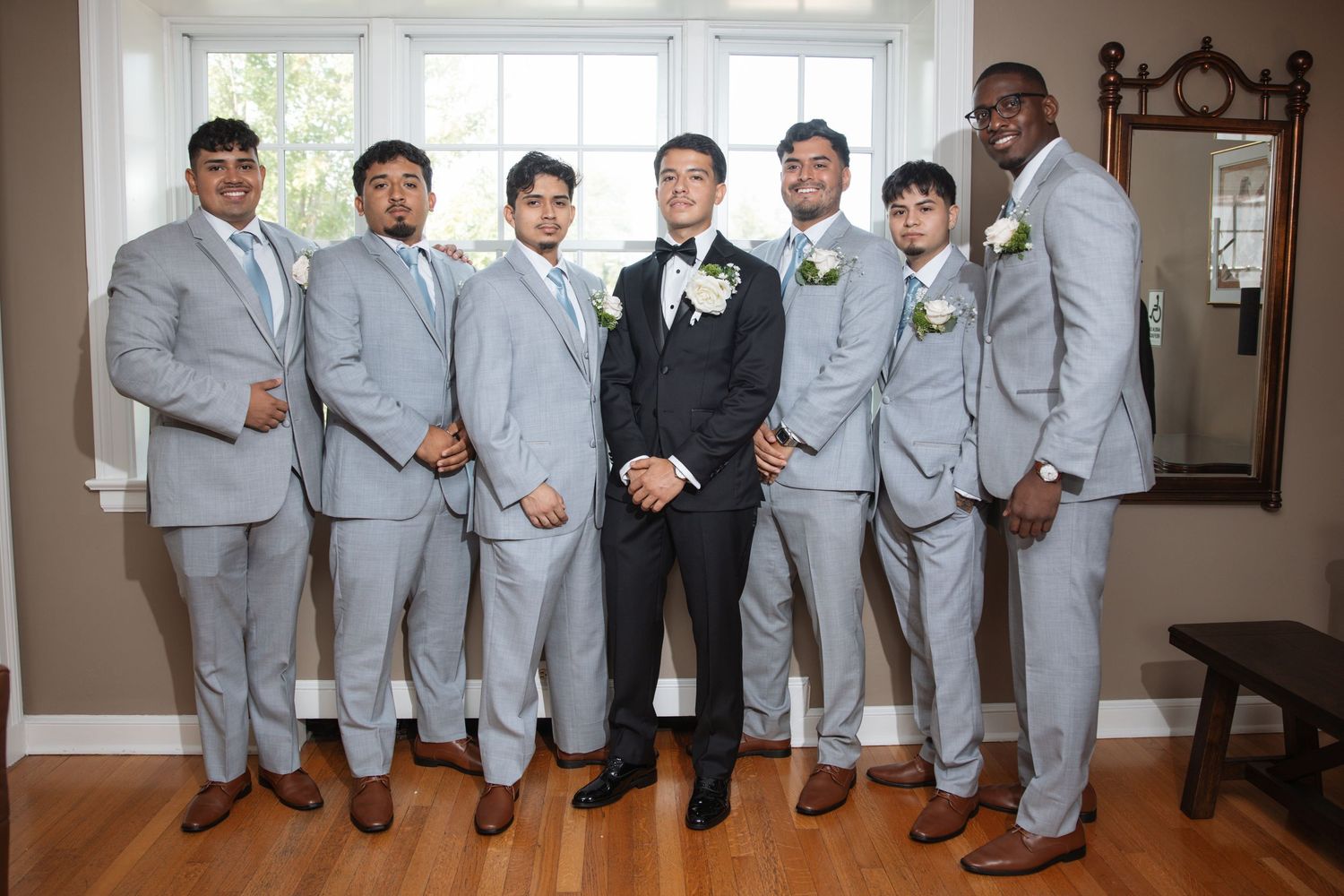 Groom surrounded by groomsmen in gray suits posing indoors before wedding.