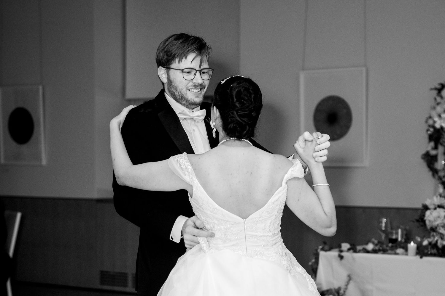 Black and white photo of smiling groom dancing with bride in off-shoulder gown at wedding reception.