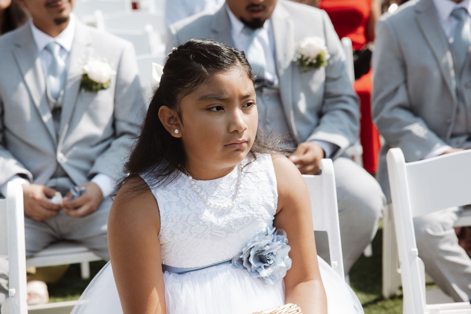Young flower girl in white dress with blue floral sash sits at outdoor wedding ceremony.
