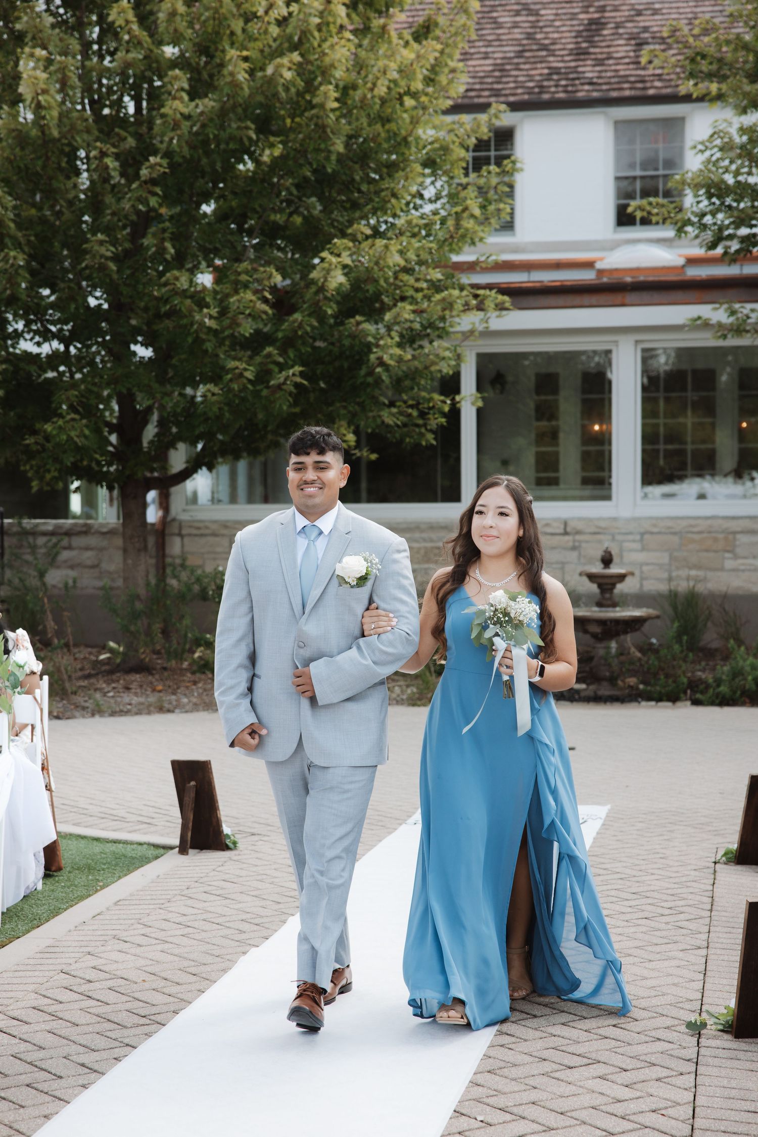 Smiling bridesmaid in blue dress walks with groomsman in gray suit down wedding aisle.