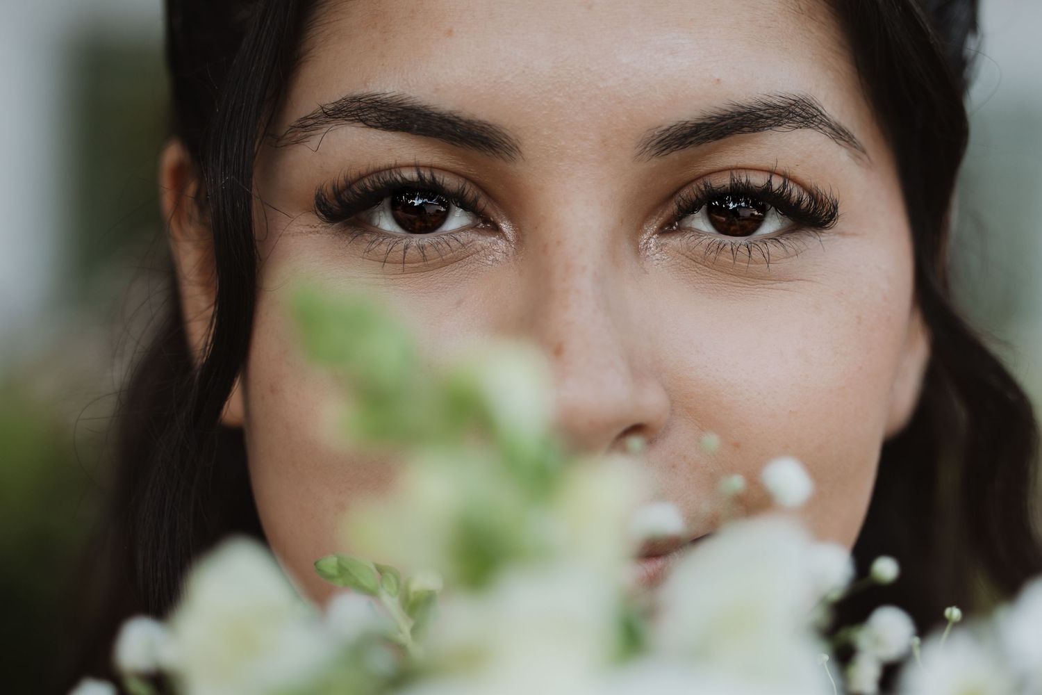 Close-up portrait of bride with dark eyes and lashes, partially obscured by white wedding bouquet flowers.