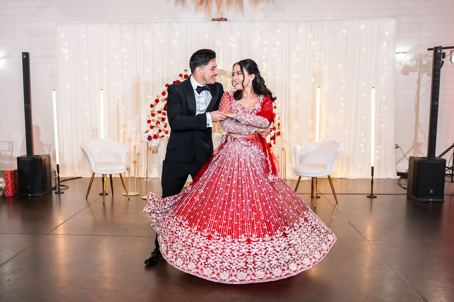 Indian bride in ornate red lehenga spins elegantly with groom in black tuxedo during wedding first dance.