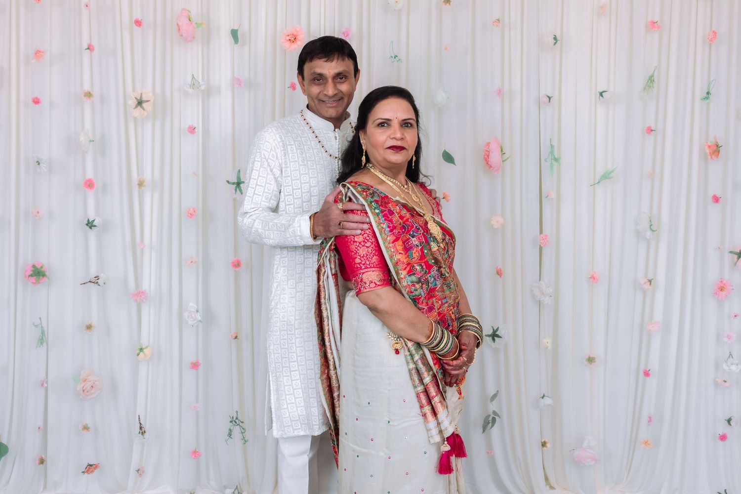 Indian couple posing in traditional wedding attire, man in white sherwani and woman in pink and white saree.