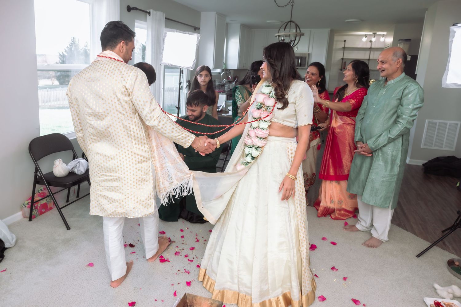Indian bride and groom in traditional white attire hold hands during wedding ceremony with family watching.