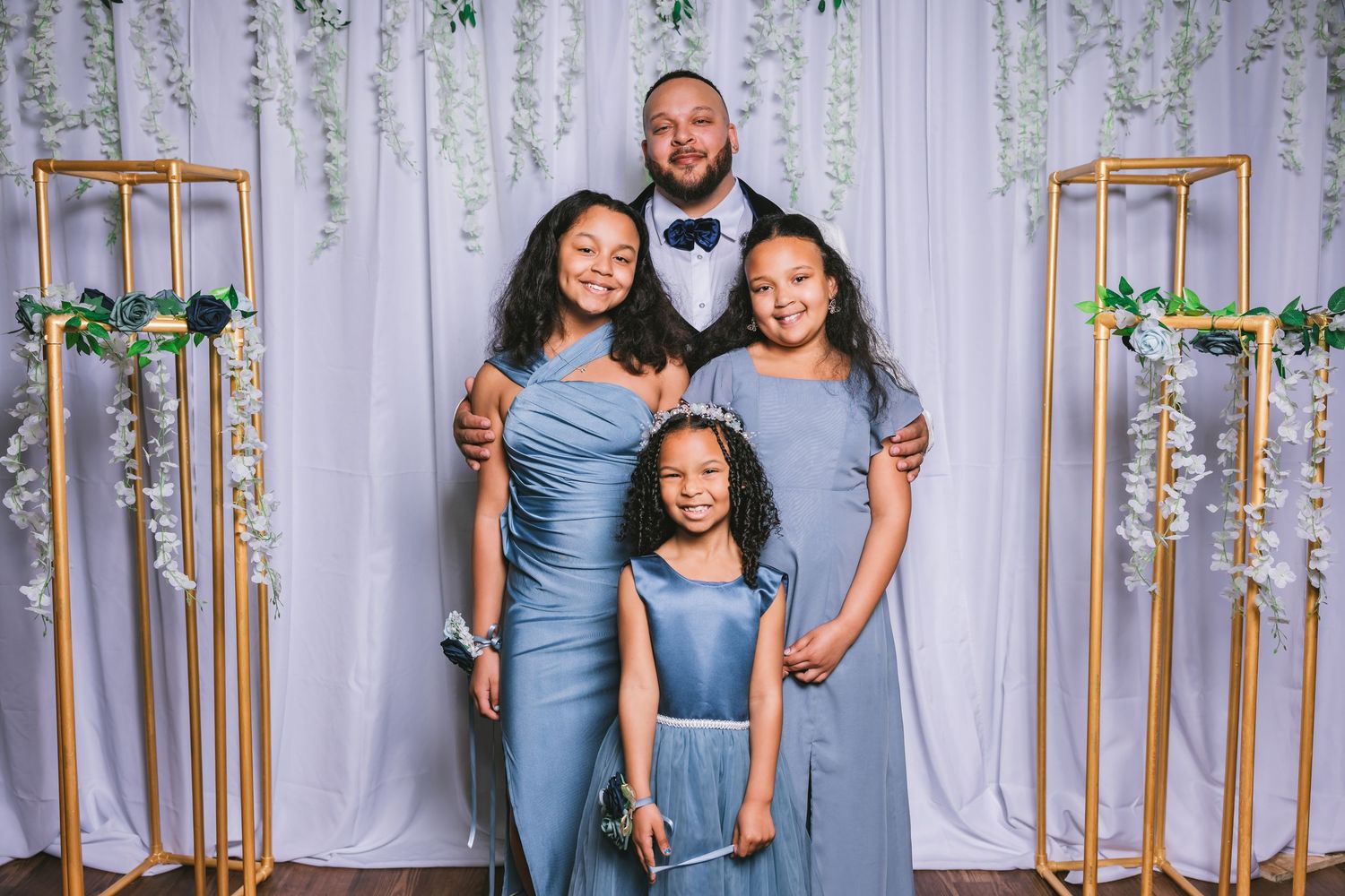 Groom posing with three girls in dusty blue dresses in front of white floral backdrop.