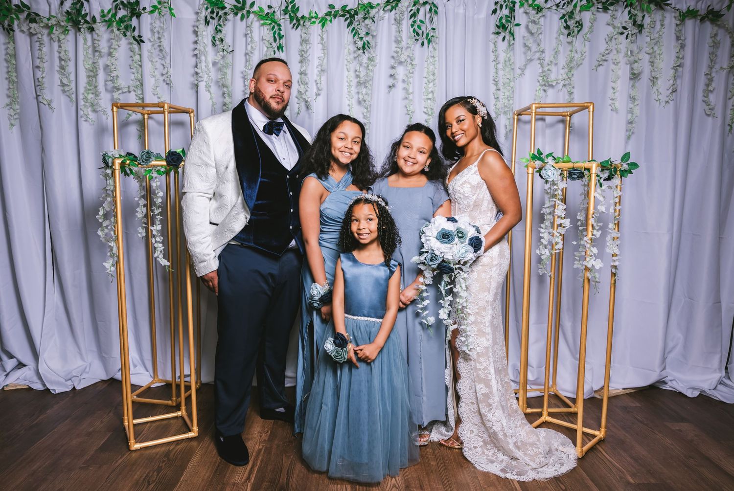 Wedding family portrait with groom in white tuxedo, bride in lace gown, and three girls in blue dresses before floral backdrop.
