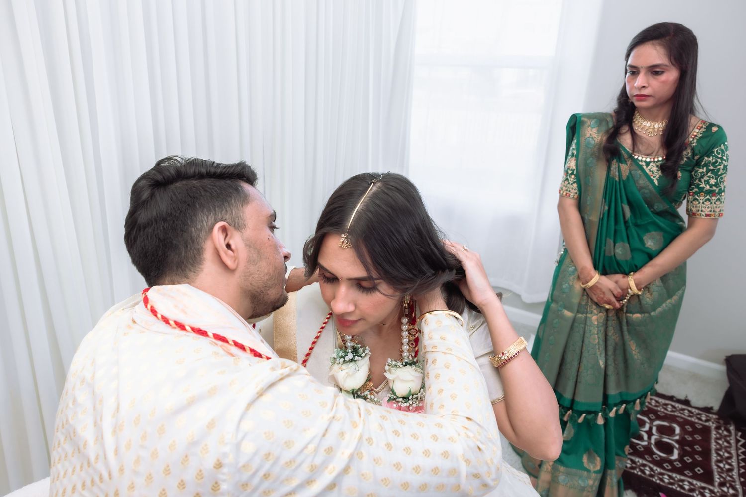 Groom places maang tikka on bride while woman in green saree watches Hindu wedding ceremony.