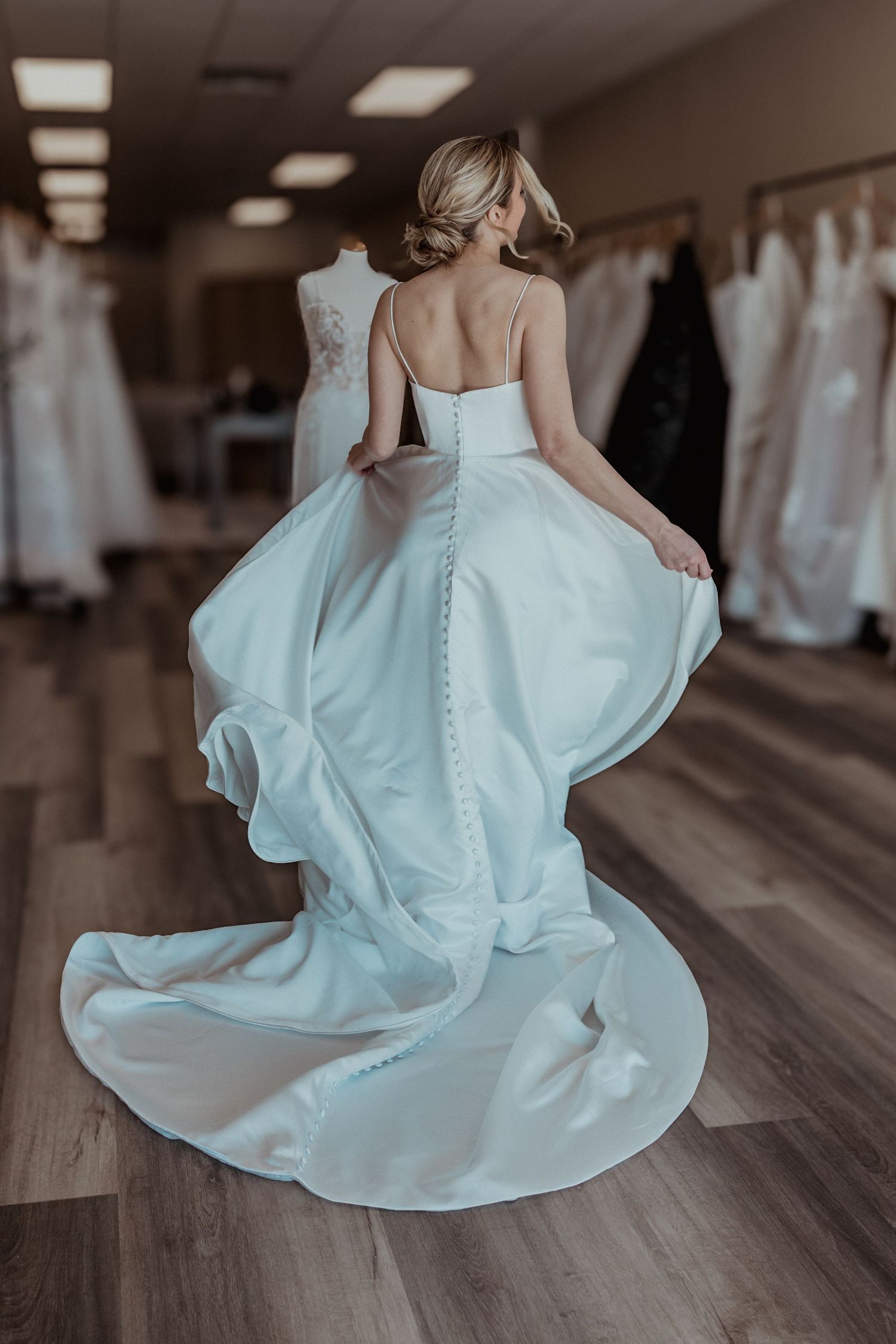 Bride from behind spinning in white satin wedding gown with button detail train in bridal boutique.