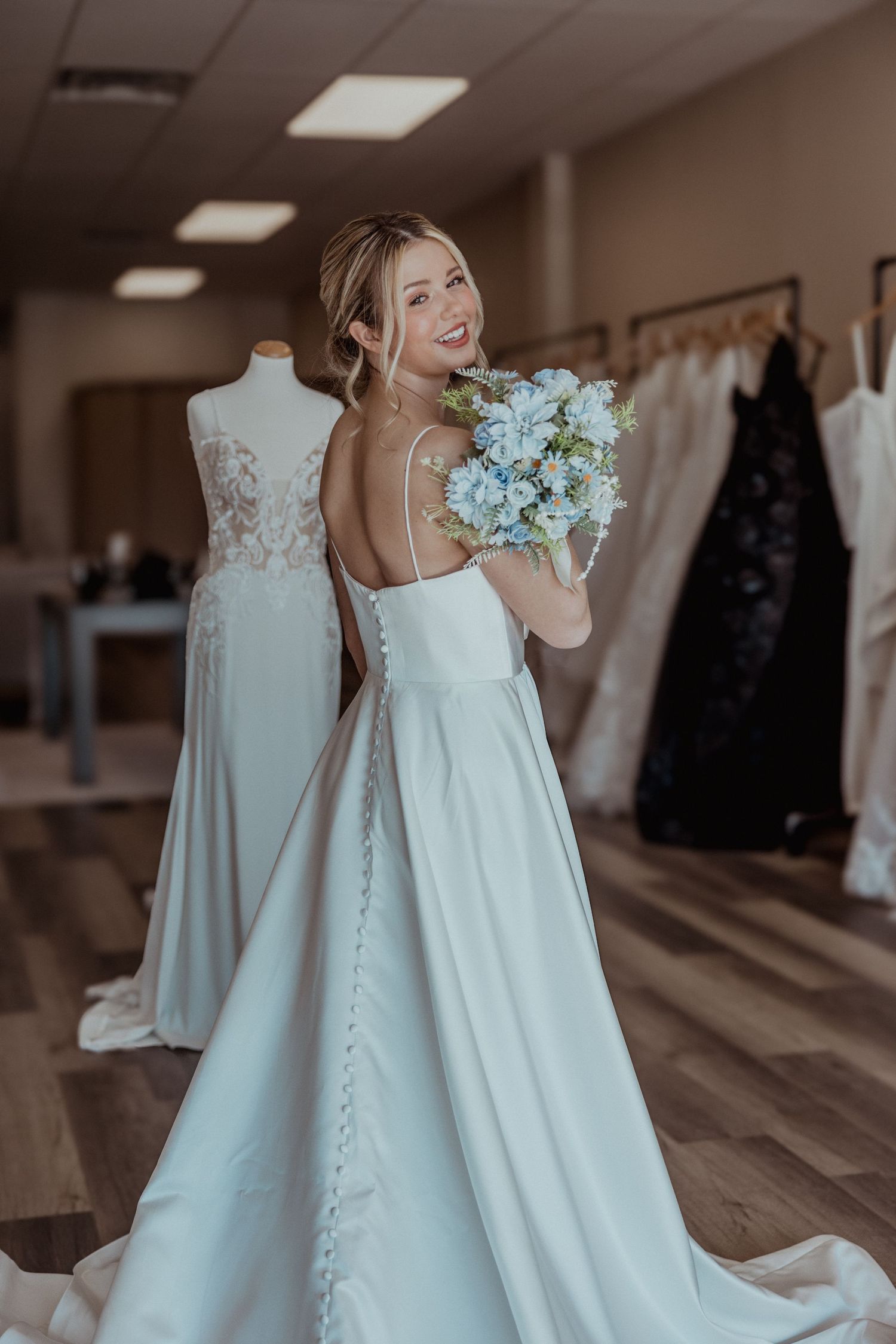 Smiling bride twirling in sleek white satin backless wedding dress holding blue floral bouquet in bridal shop.