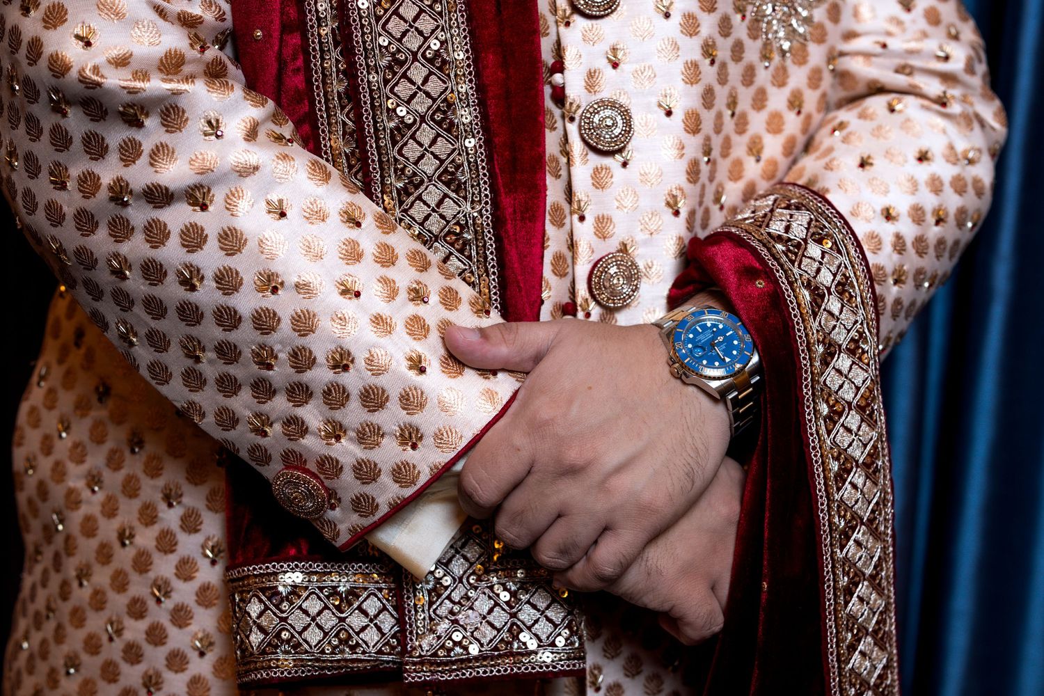 Close-up of Indian groom in gold sherwani with blue watch and red embroidered dupatta detail.