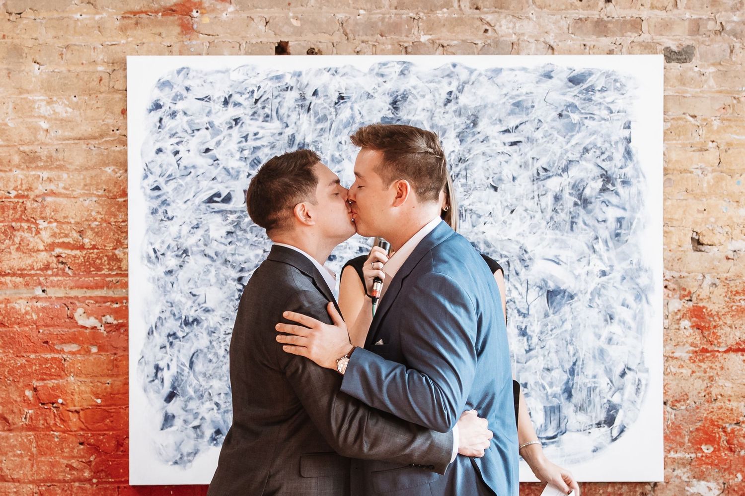 Two grooms share a first kiss in front of a large blue and white abstract painting at their wedding.