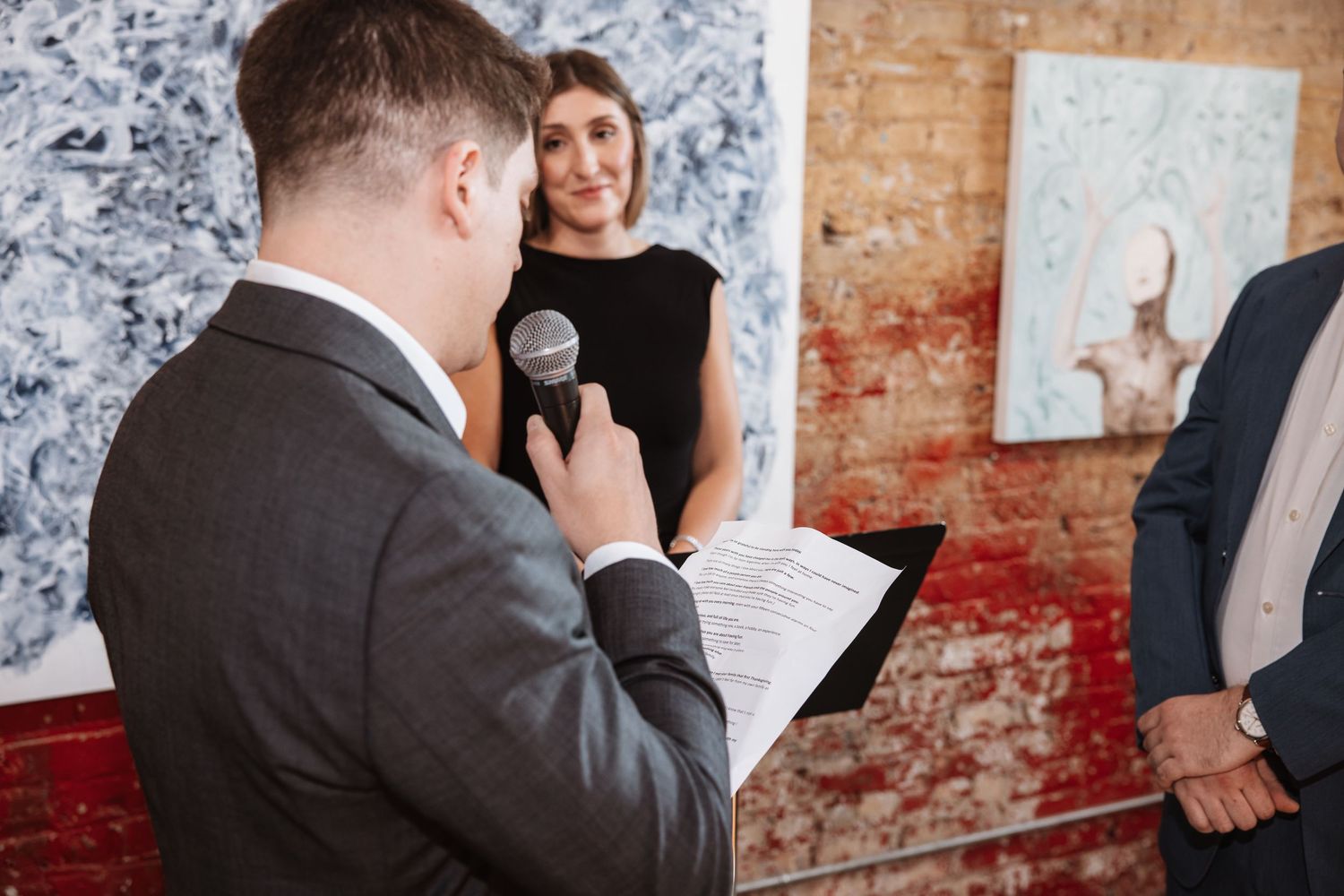 Officiant reads from clipboard while conducting wedding ceremony in art gallery venue.