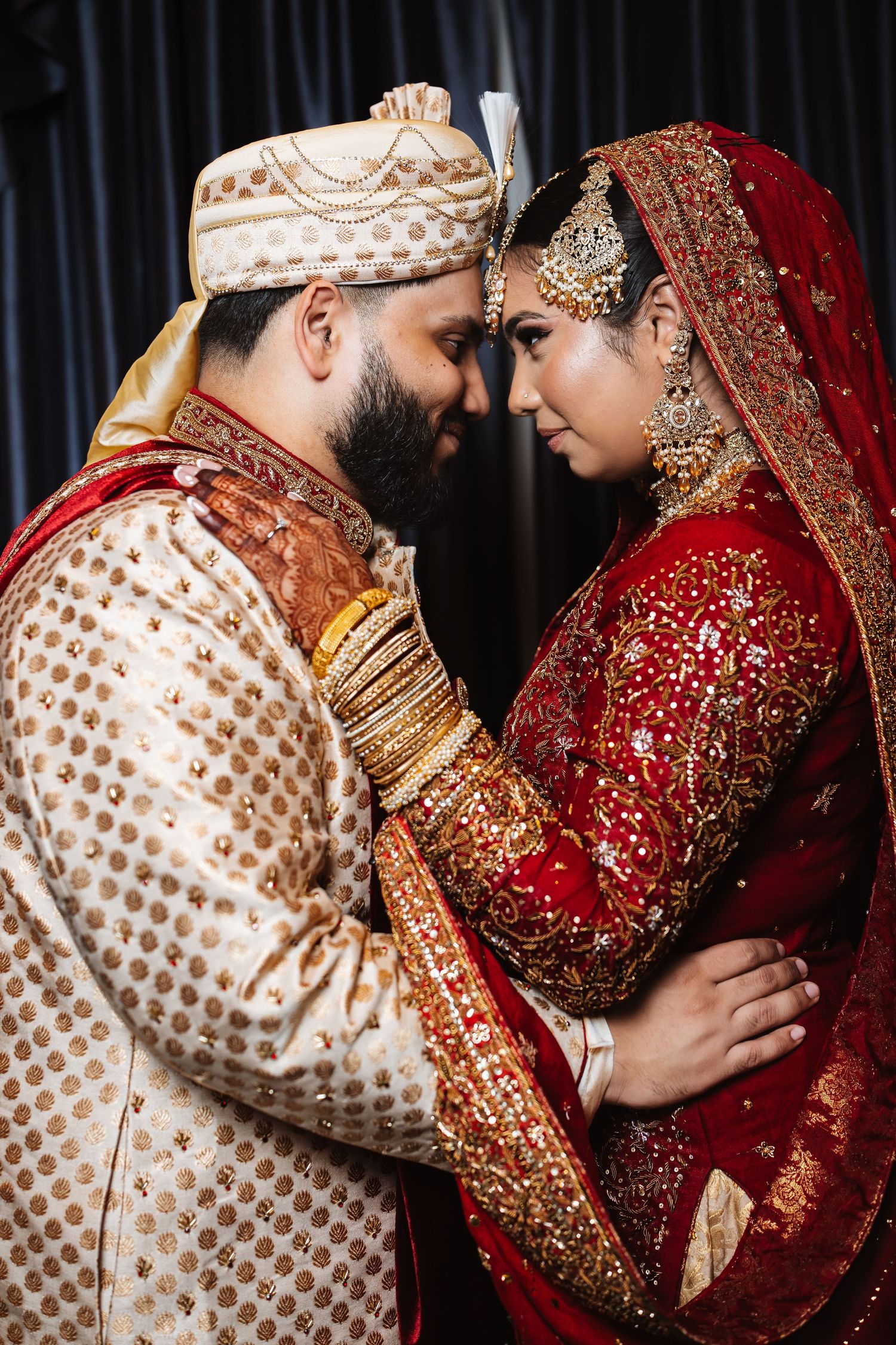 Indian bride in red lehenga and groom in gold sherwani sharing a romantic forehead-touch pose.