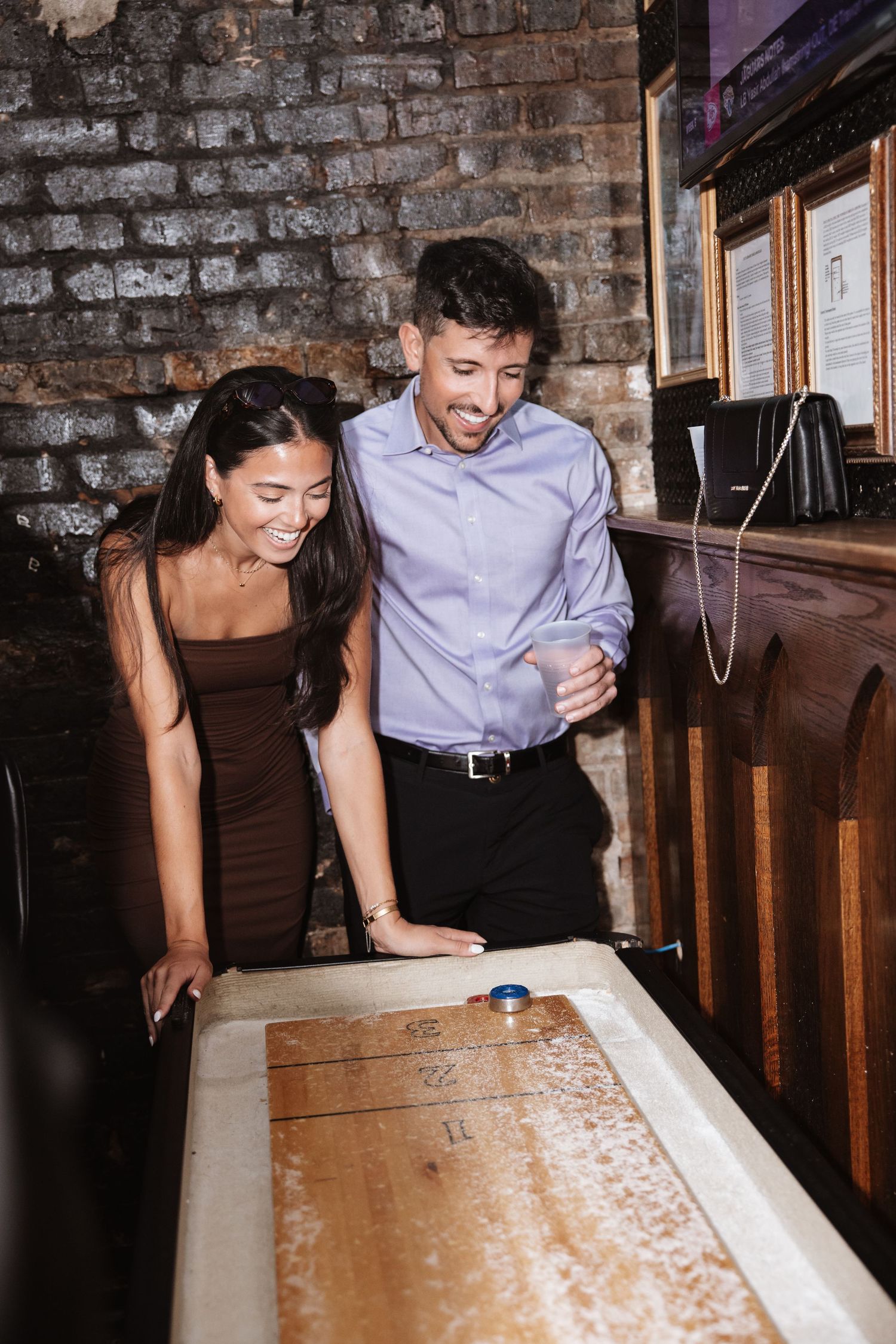 Couple smiling while playing shuffleboard together at a rustic brick-walled bar.