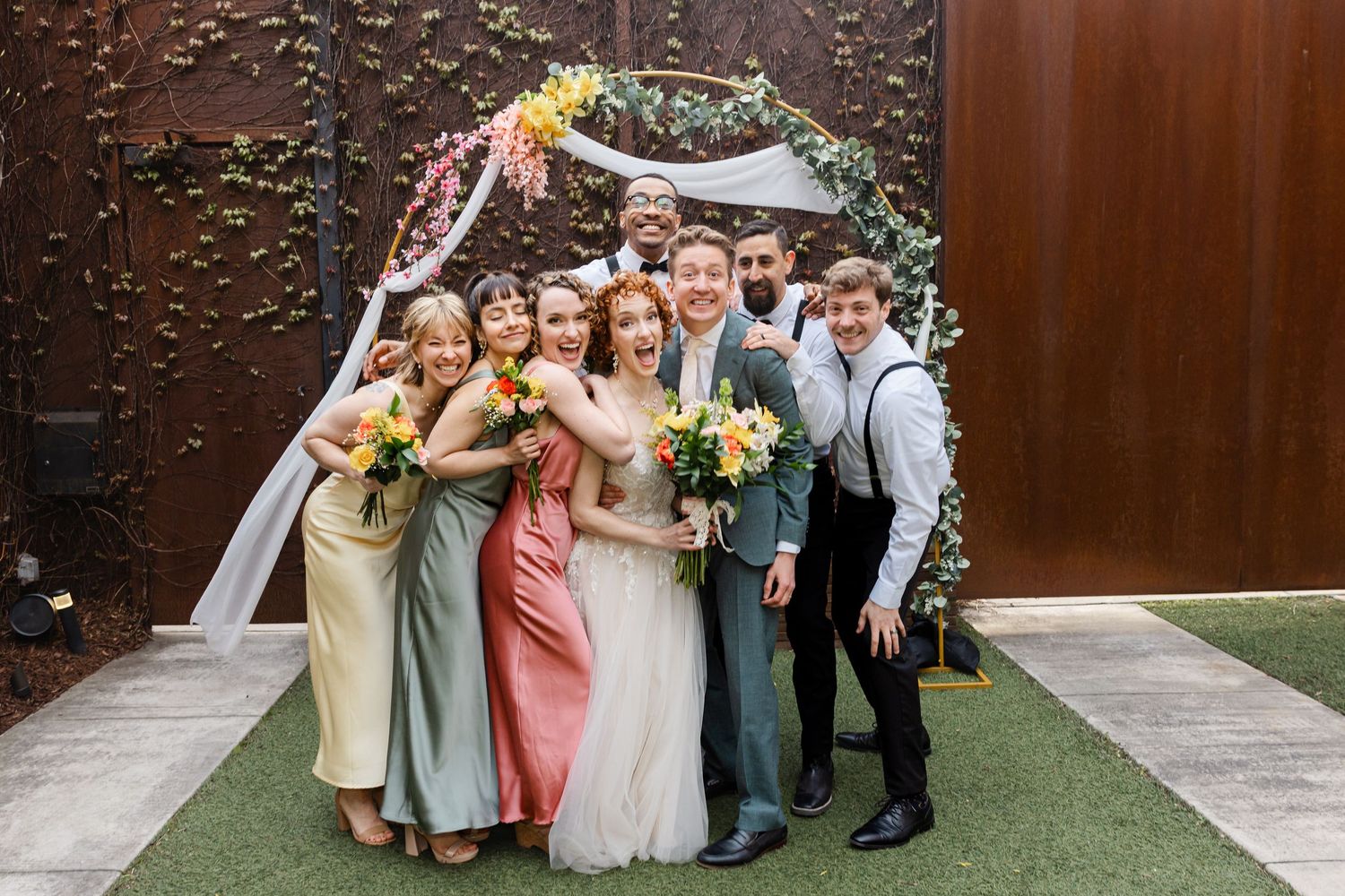 Bridal party laughs and huddles together for a fun group photo in front of a decorated floral wedding arch.