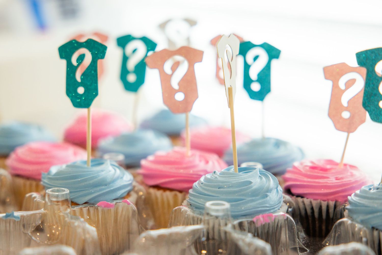 Gender reveal cupcakes with pink and blue frosting topped with baby onesie and question mark decorations.