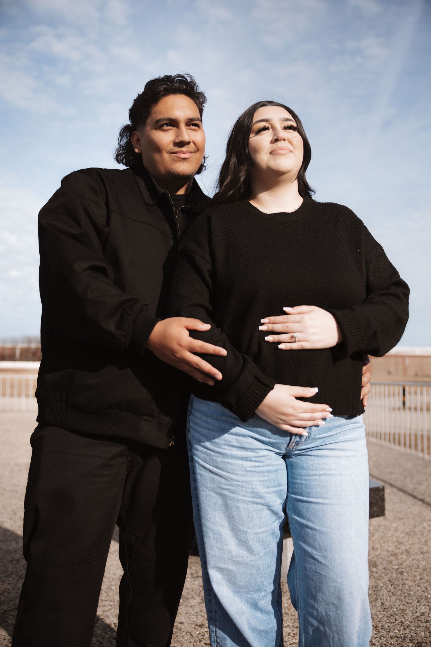 Couple cradles pregnant belly outdoors under blue sky in matching black outfits.