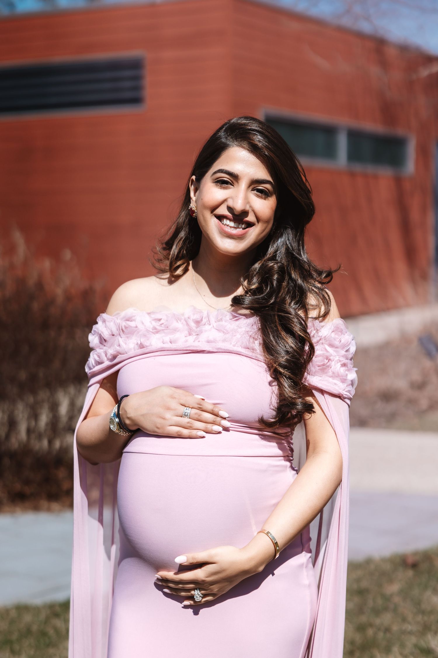 Happy pregnant woman in pink off-shoulder dress with floral details poses outdoors smiling.