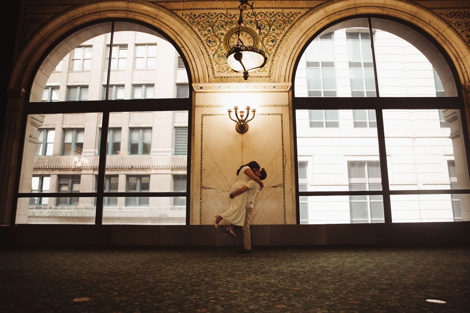Couple embraces romantically in grand hall with arched windows.