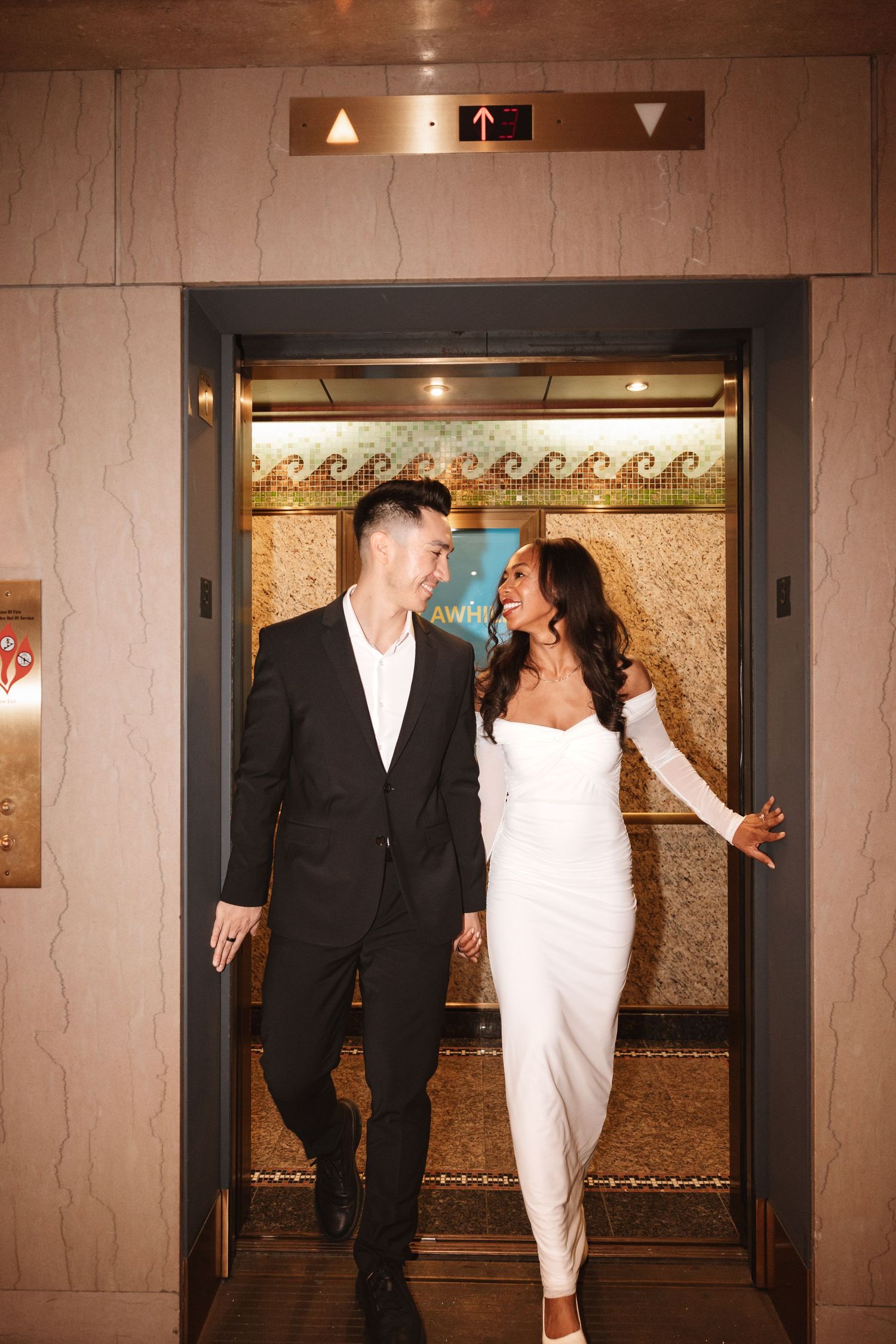 Couple laughing and holding hands exiting decorative elevator, dressed in black suit and white bridal gown.