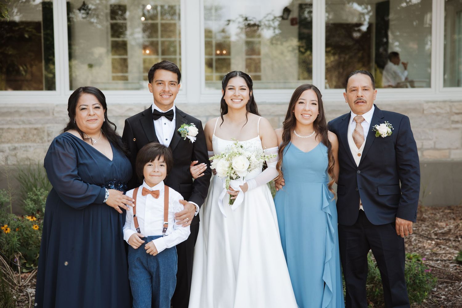 Bride and groom pose with family of six including young boy in suspenders outside wedding venue.