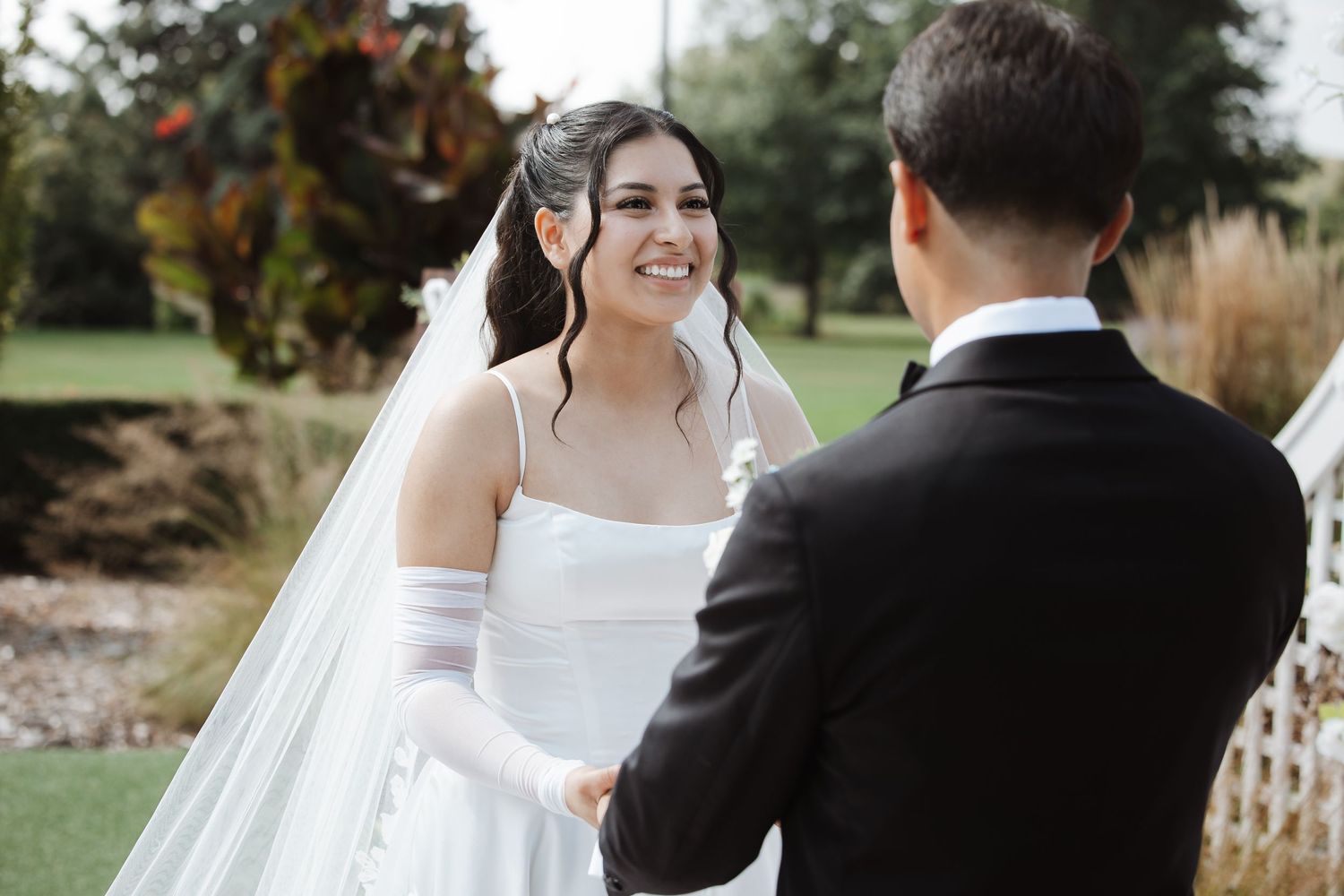 Bride in white gown and veil holds hands with groom during outdoor wedding ceremony.