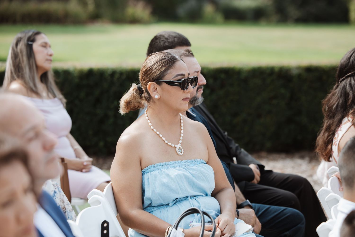 A wedding guest in a light blue dress and pearl necklace wearing sunglasses seated at an outdoor ceremony.