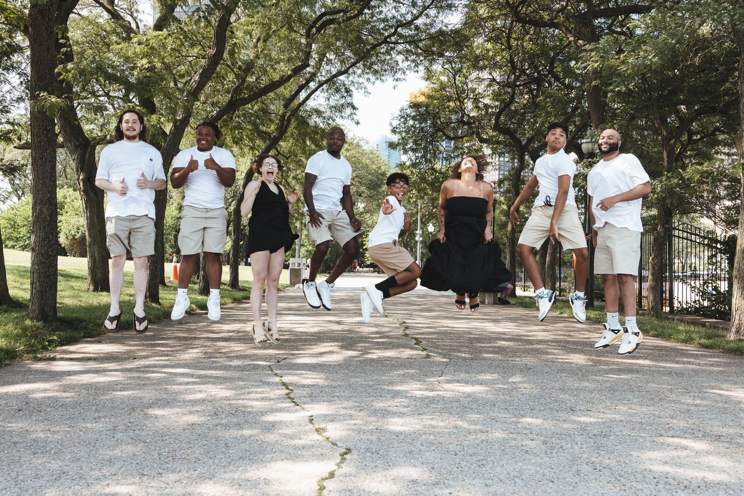 Energetic group photo of nine people jumping outdoors on a sunny tree-lined park path.