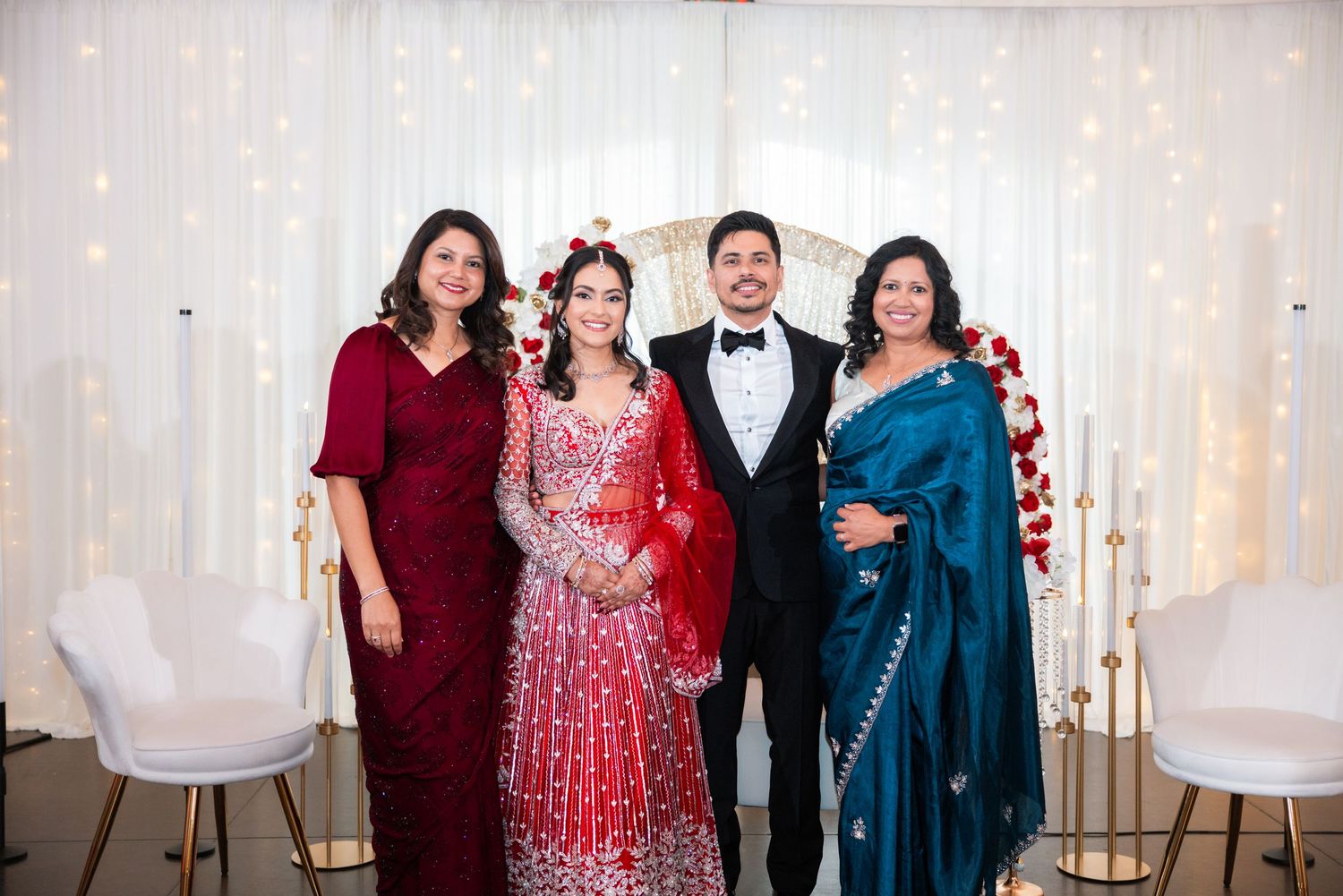 South Asian wedding photo featuring bride in embroidered red lehenga, groom in black tuxedo, posing with two family members.