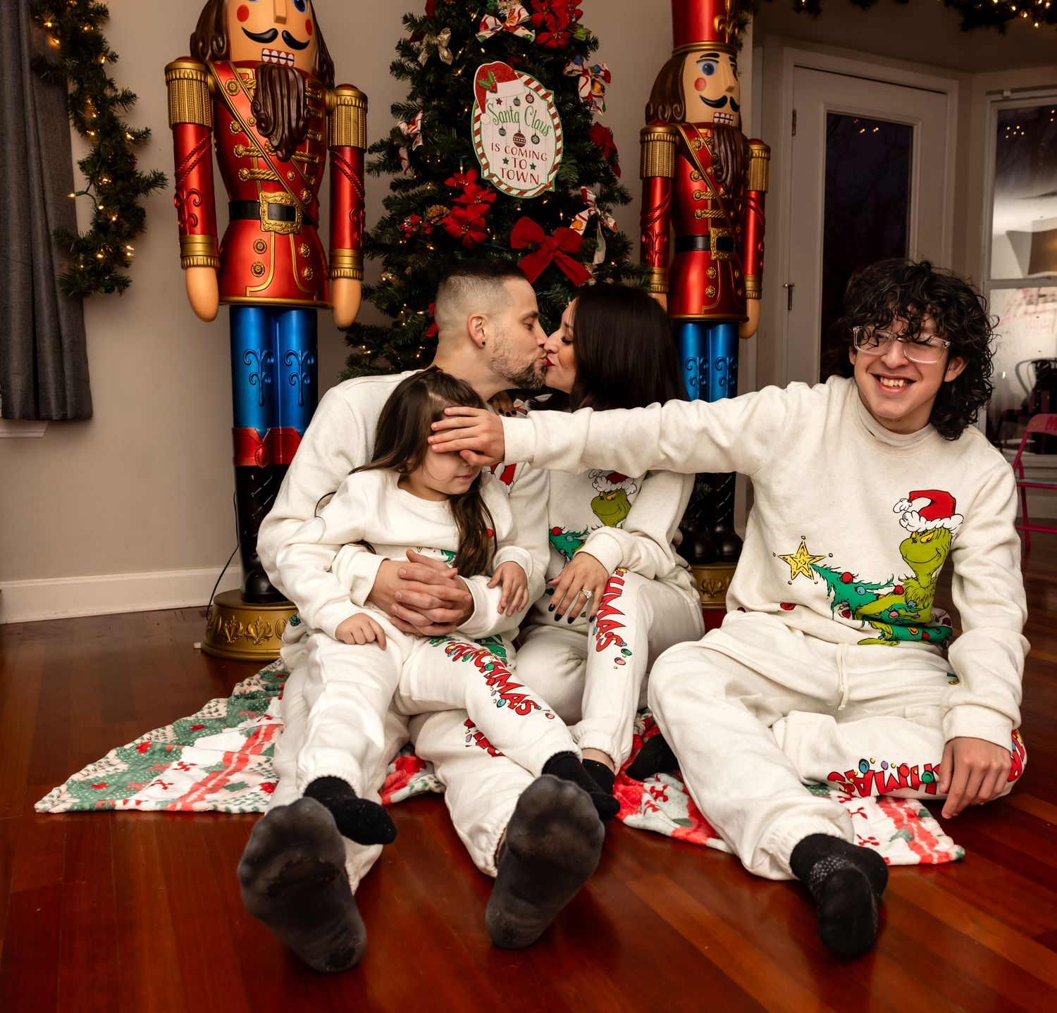 Family in matching Grinch Christmas pajamas with parents kissing near decorated holiday tree.