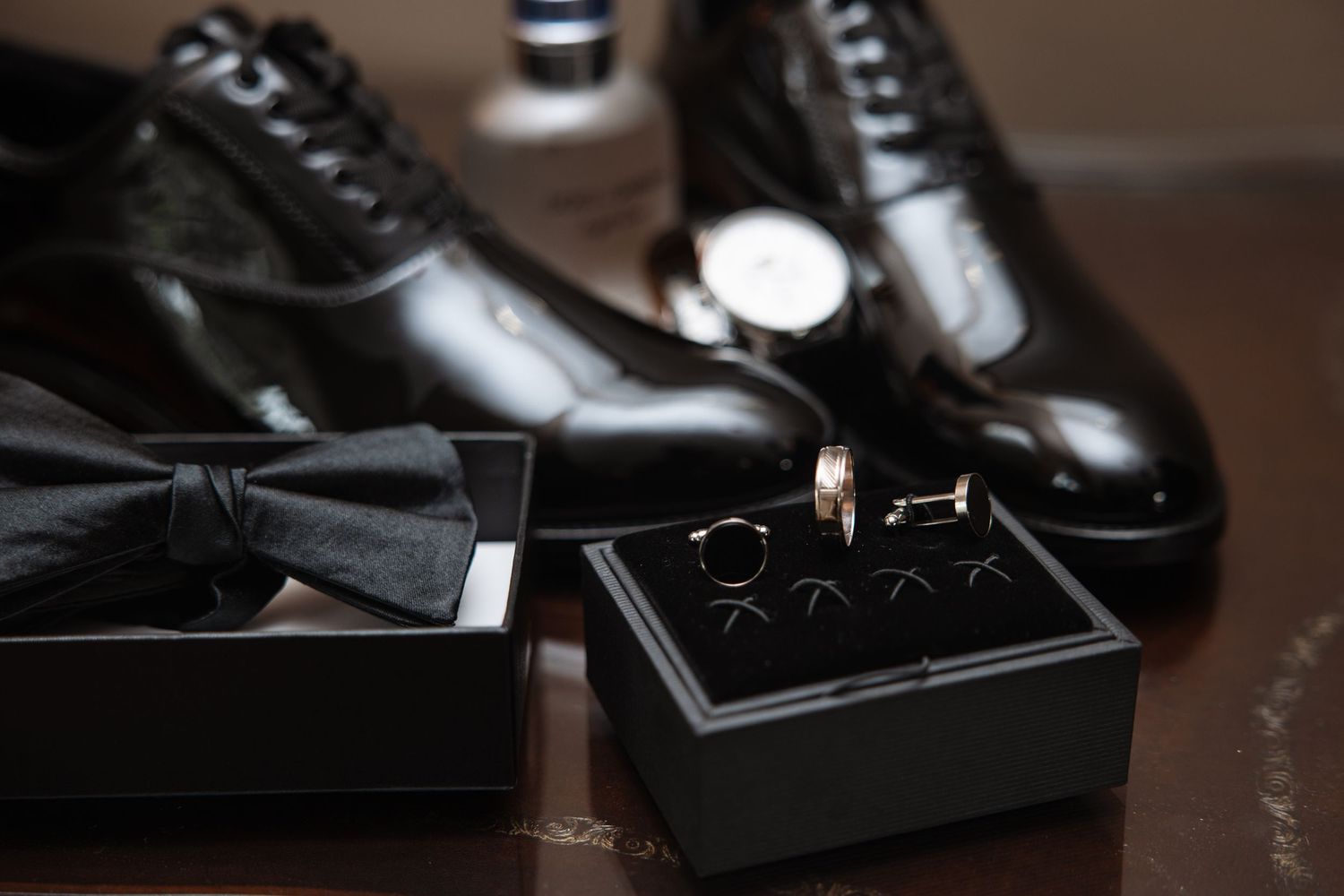 Close-up of groom accessories including black patent shoes, bow tie, cufflinks, wedding rings, and watch on dark surface.