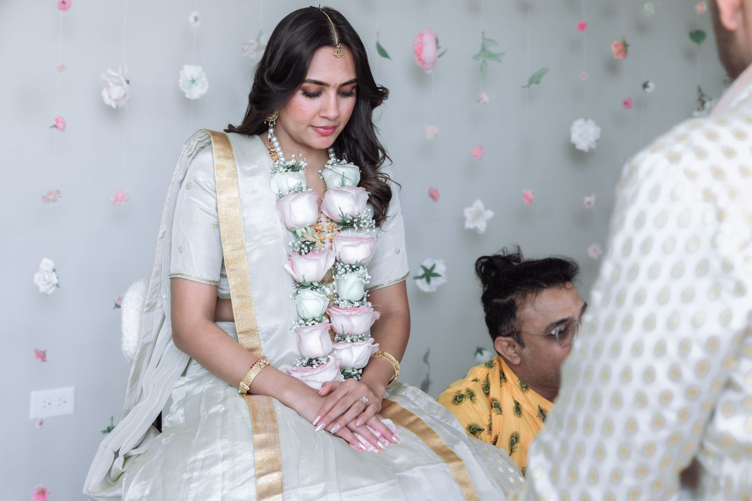 Bride in white lehenga with floral garland bows her head during an Indian wedding ceremony.
