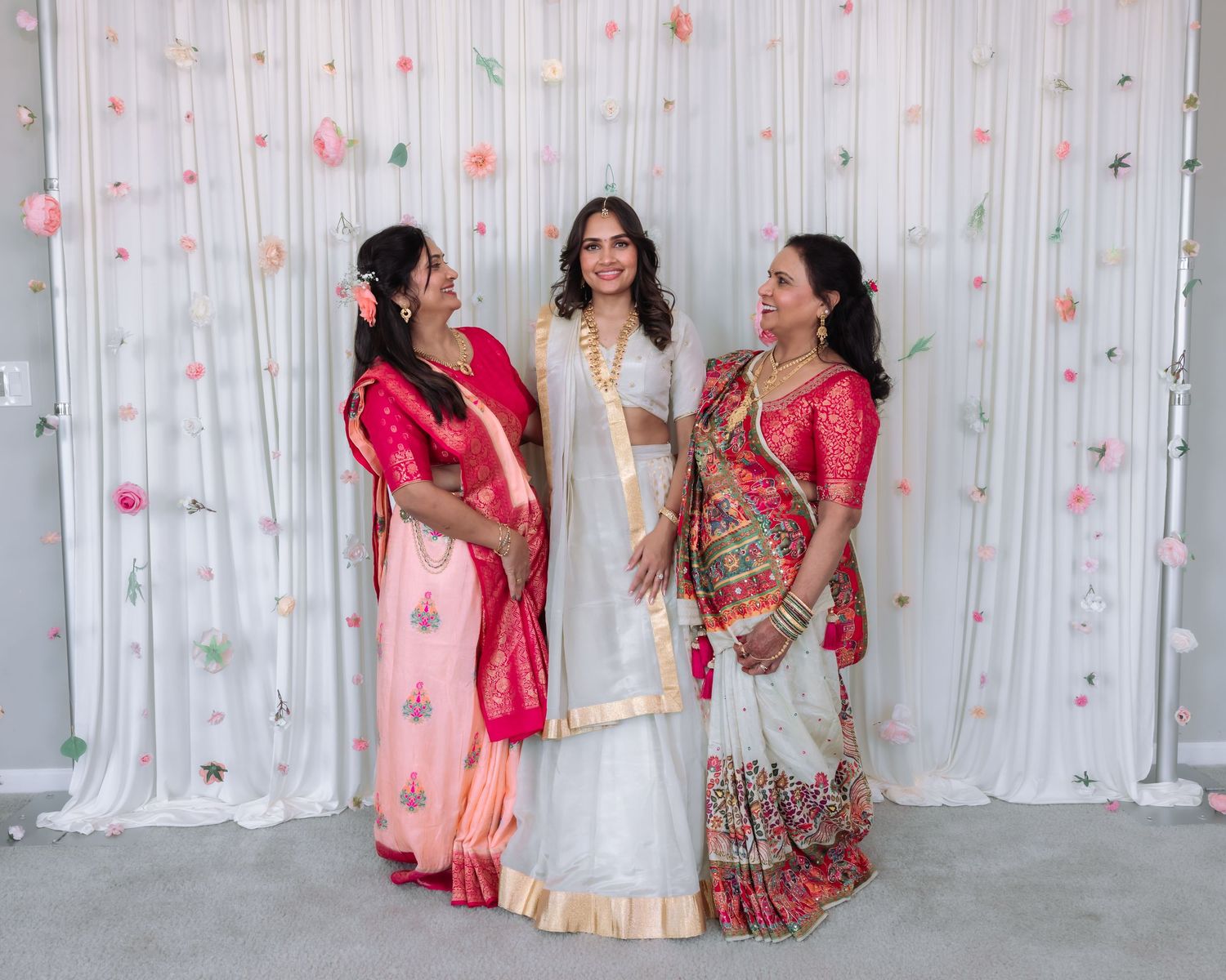 Bride in white lehenga stands between two women in pink and printed sarees at an Indian wedding celebration.