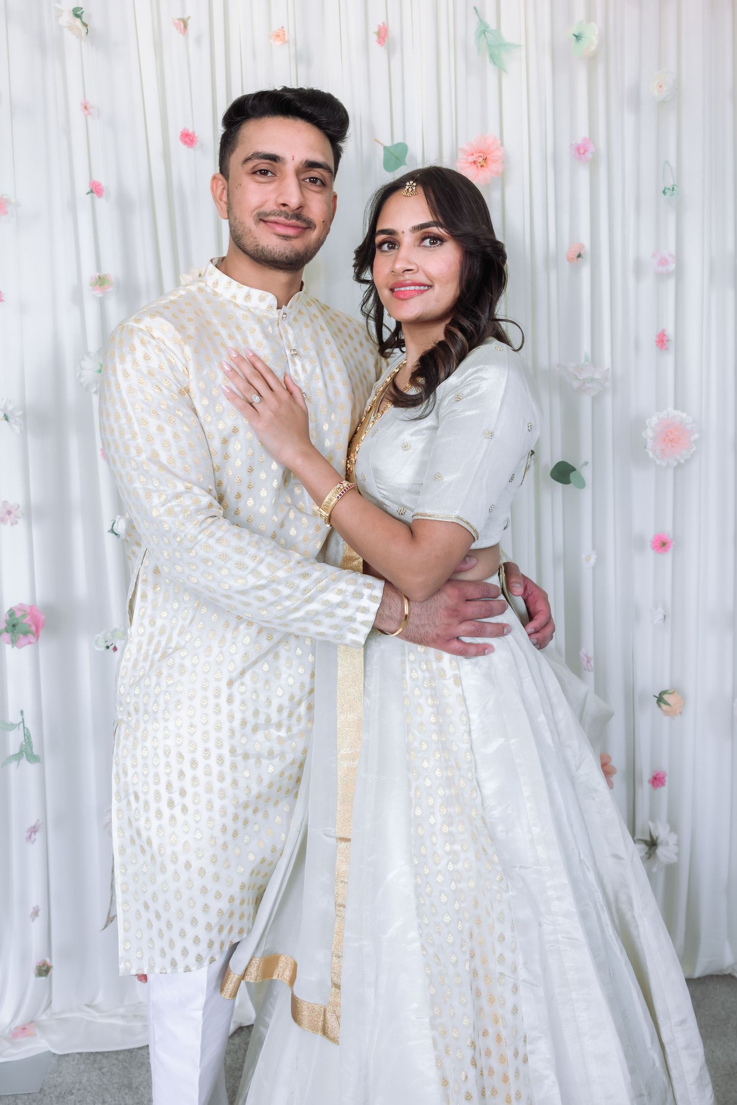 Indian couple in coordinating white and gold traditional outfits embracing at their engagement celebration.