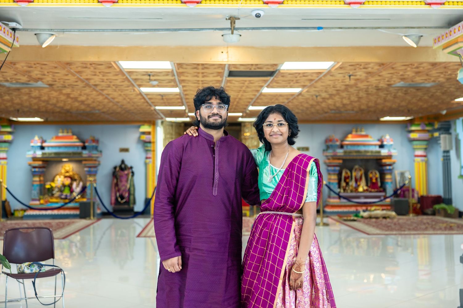 Young couple in traditional Indian attire smiling together inside a decorated Hindu temple.