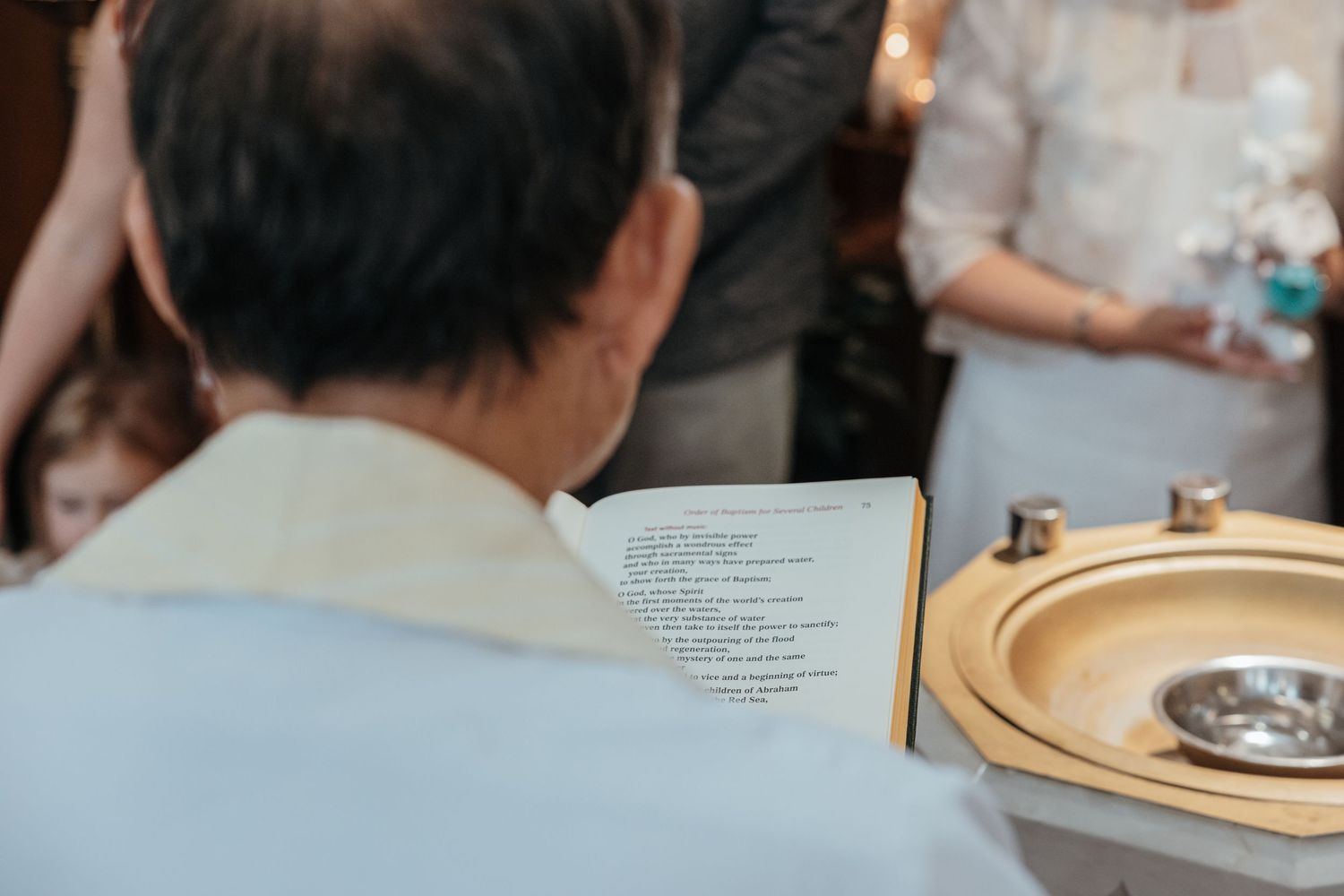 A Catholic priest reads baptismal rites from a book positioned beside a golden baptismal font.