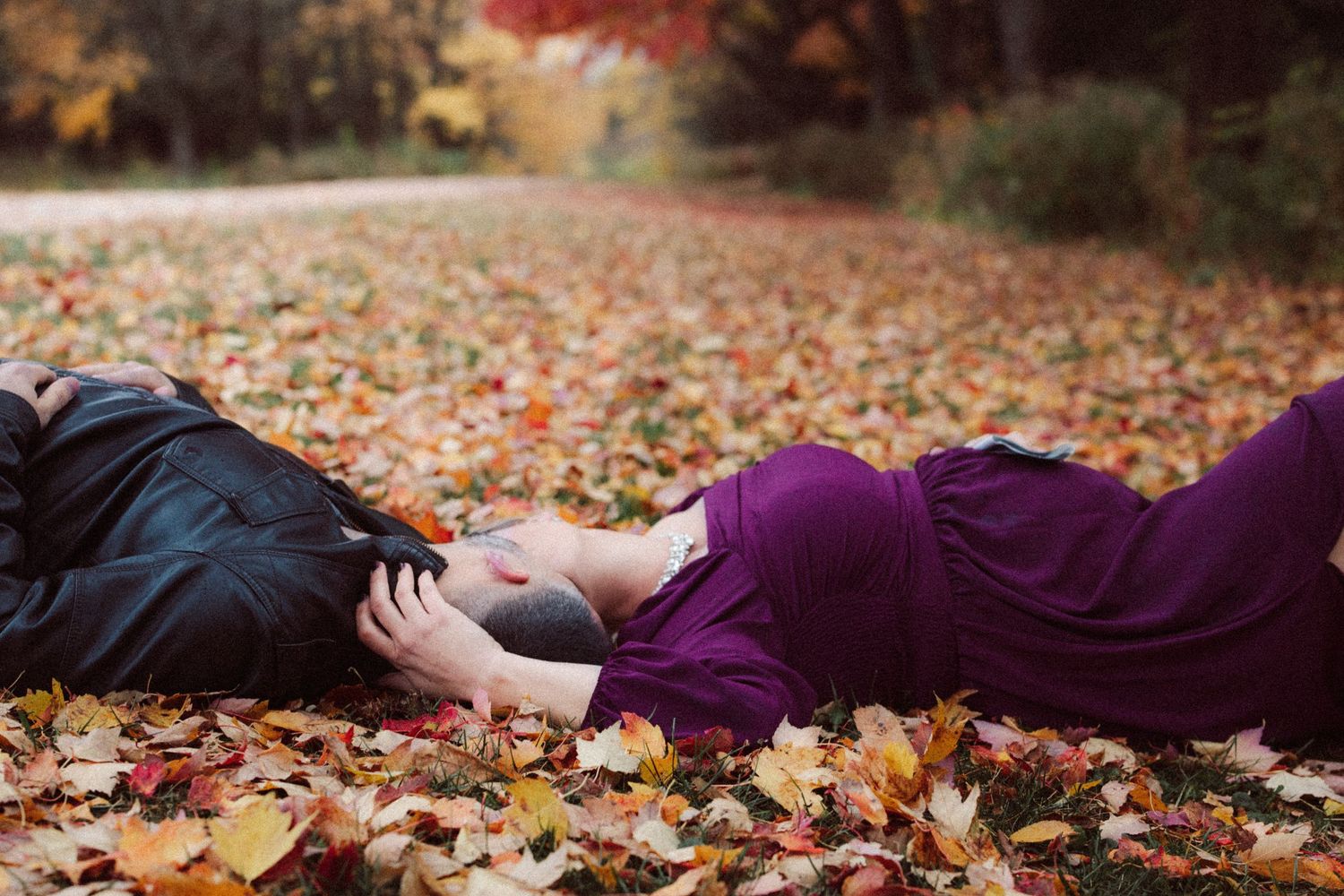 Couple lying among colorful autumn leaves, pregnant woman in purple dress resting beside her partner outdoors.