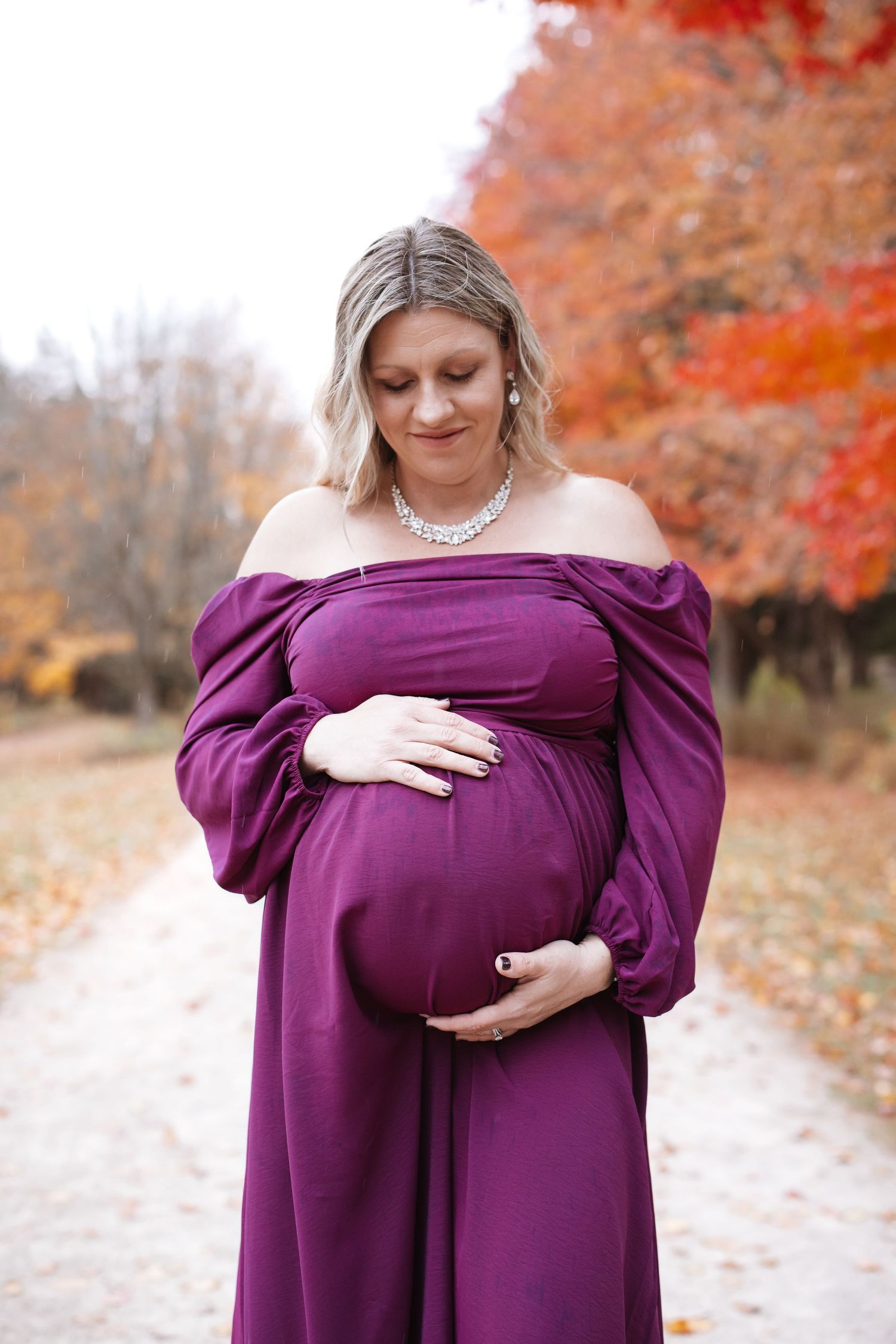 Pregnant woman in purple off-shoulder gown and pearl necklace cradling bump on autumn pathway.