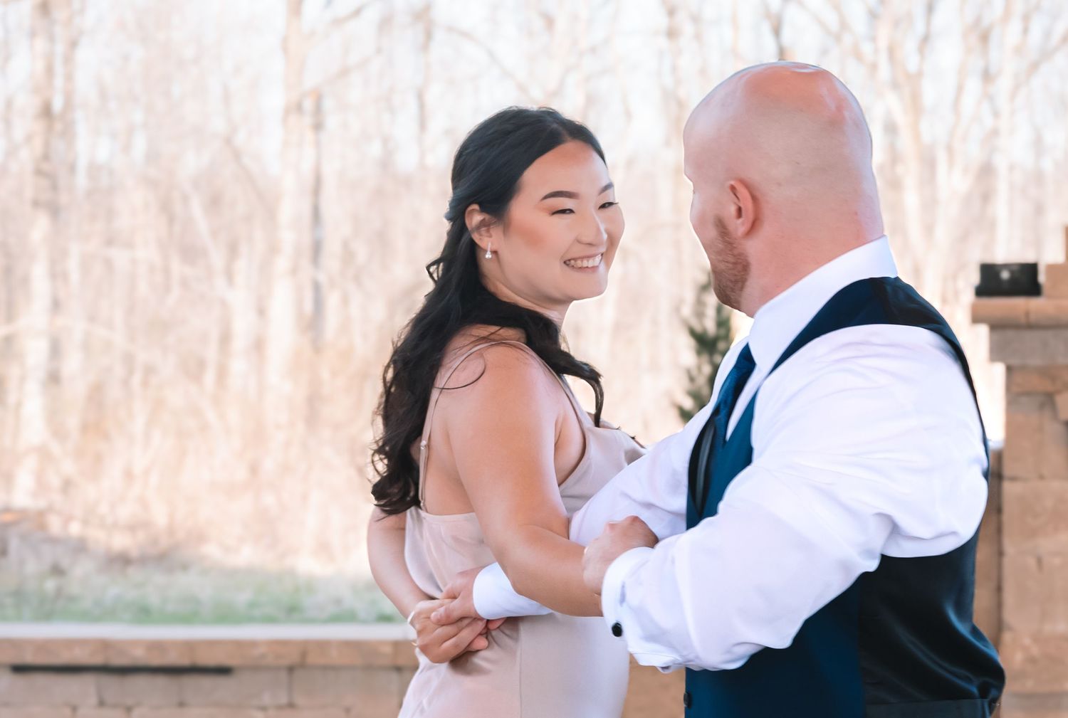 Smiling bride in blush gown dances with bald groom in navy vest at outdoor wedding reception.