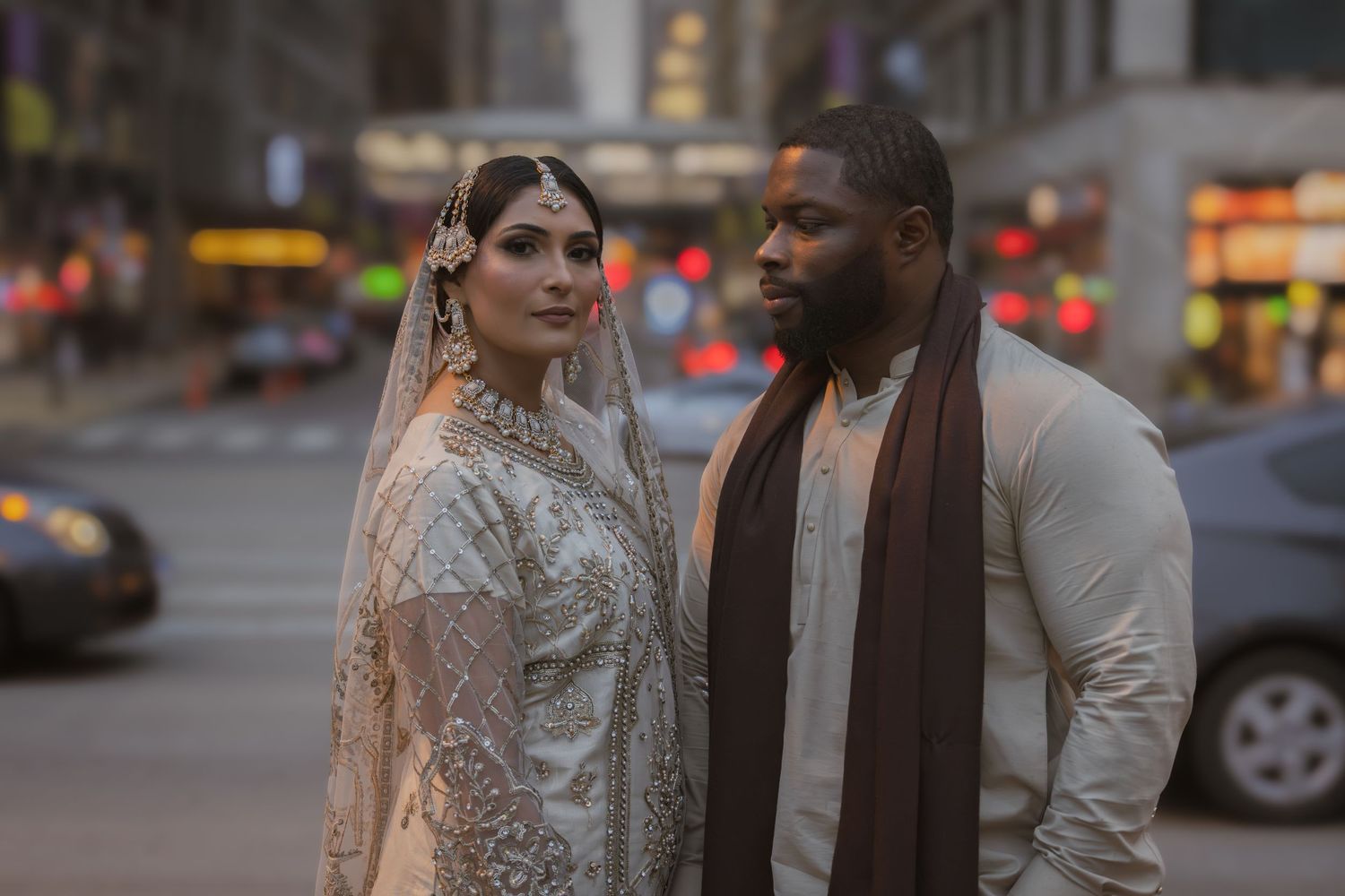 Bride in white embroidered lehenga and groom in cream kurta pose on a busy city street at dusk.