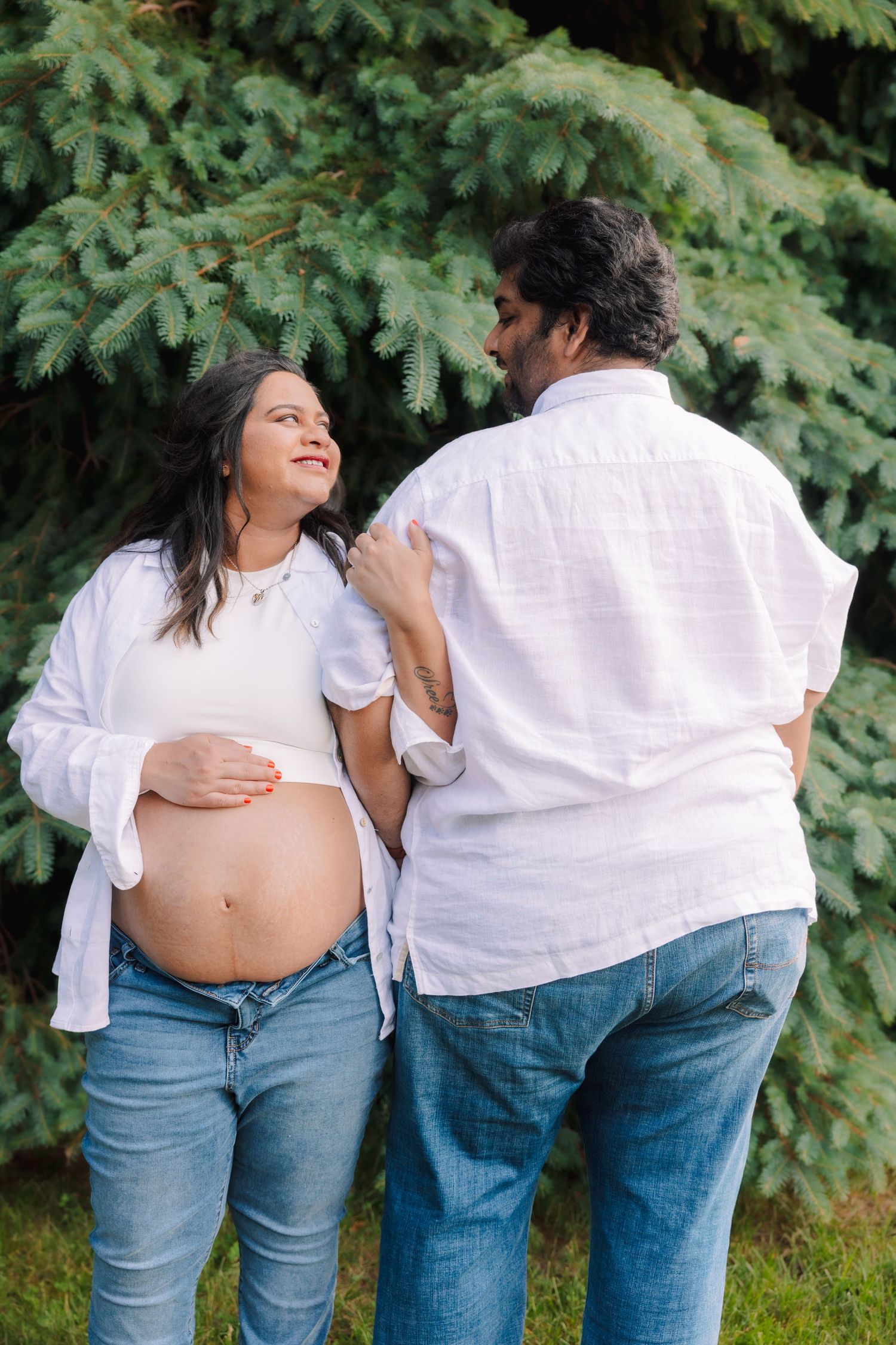 Smiling pregnant woman in white shirt and jeans shares tender moment with partner outdoors.