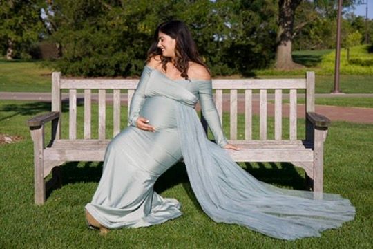 Pregnant woman in flowing sage green gown sitting gracefully on a wooden park bench on green grass.